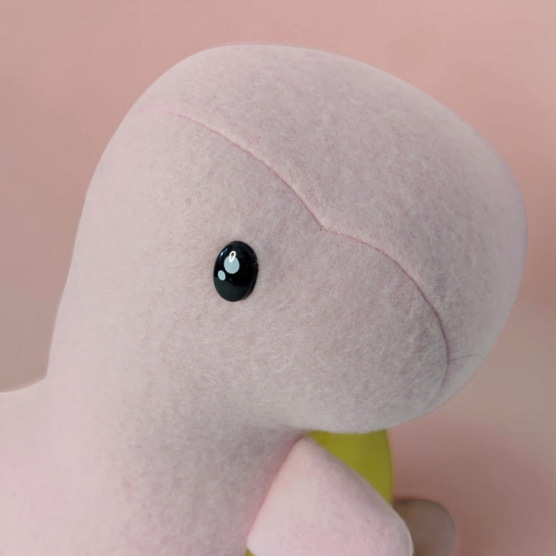 A close-up of a soft pink plush dinosaur toy with a black safety eye and a small yellow belly, set against a solid pale pink background.