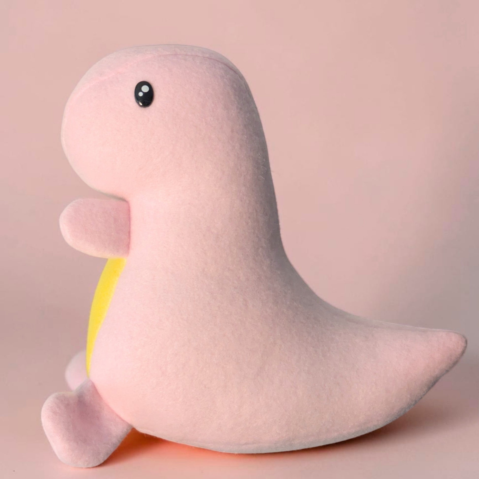 A cute, soft handmade plush toy shaped like a stylized dinosaur, in light pink with a small yellow accent on its belly and  black safety eyes, set against a solid pale pink background.