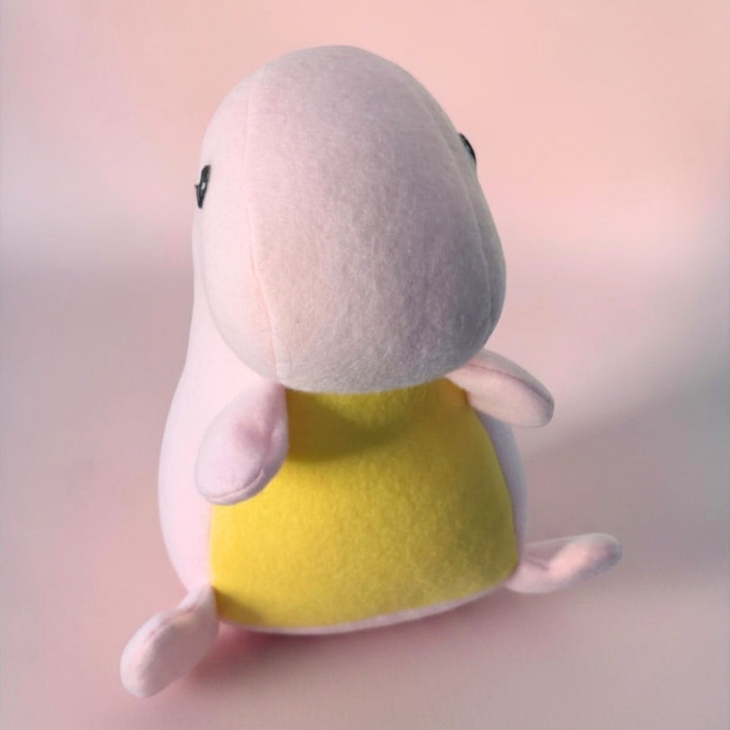 A small plush toy shaped like a rounded dinosaur character, shown from the front. The plush has a pale pink body, a bright yellow belly, and small black eyes, photographed against a soft pink studio background.