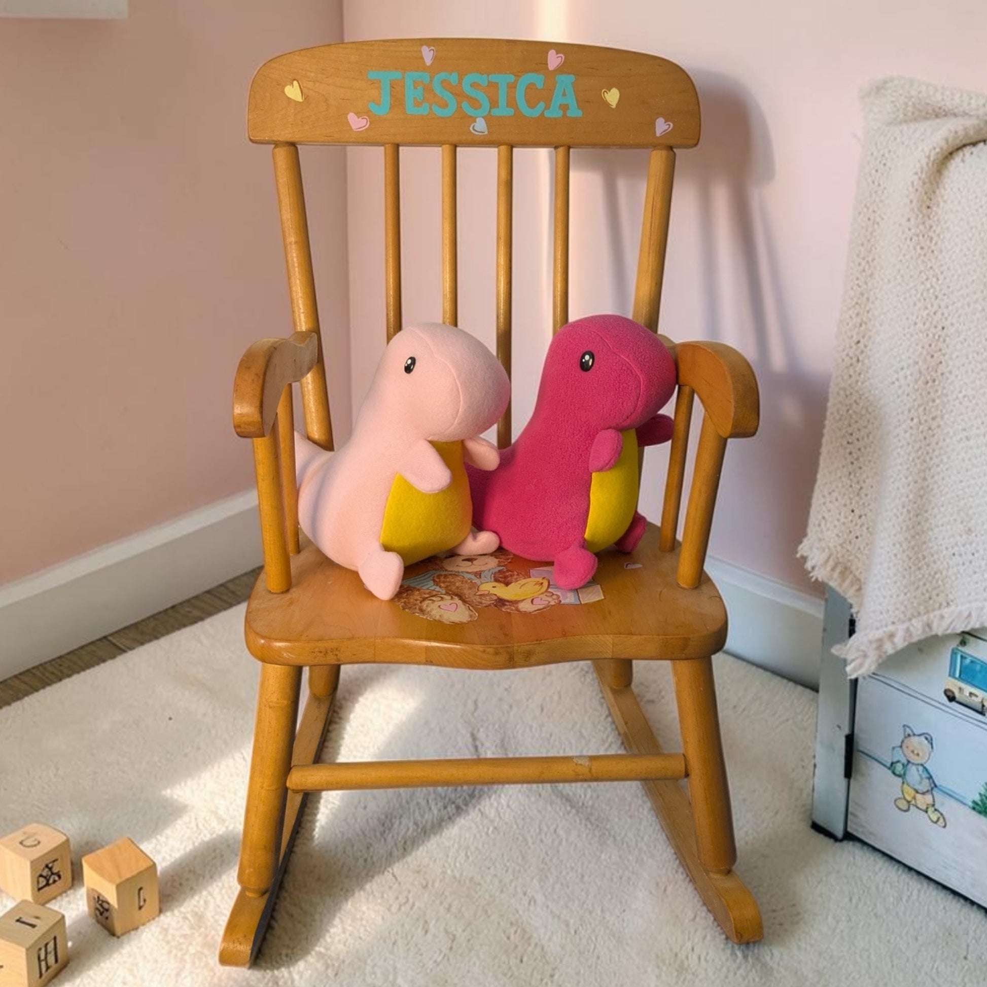 A wooden rocking chair with the name 'JESSICA' painted on the backrest, styled with two handmade plush Pink dinosaur toys on a cream carpet in a nursery setting.