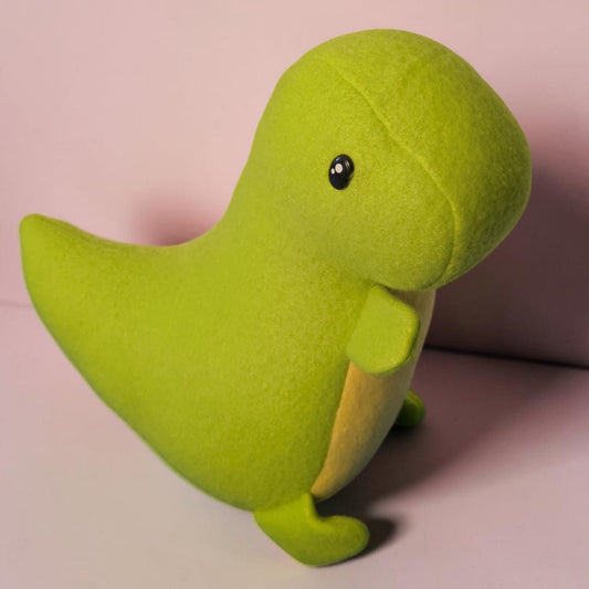 A lime green fleece handmade dinosaur plush toy with a small black safety eye and a pale yellow underside, standing on a light pink surface against a matching pink background.