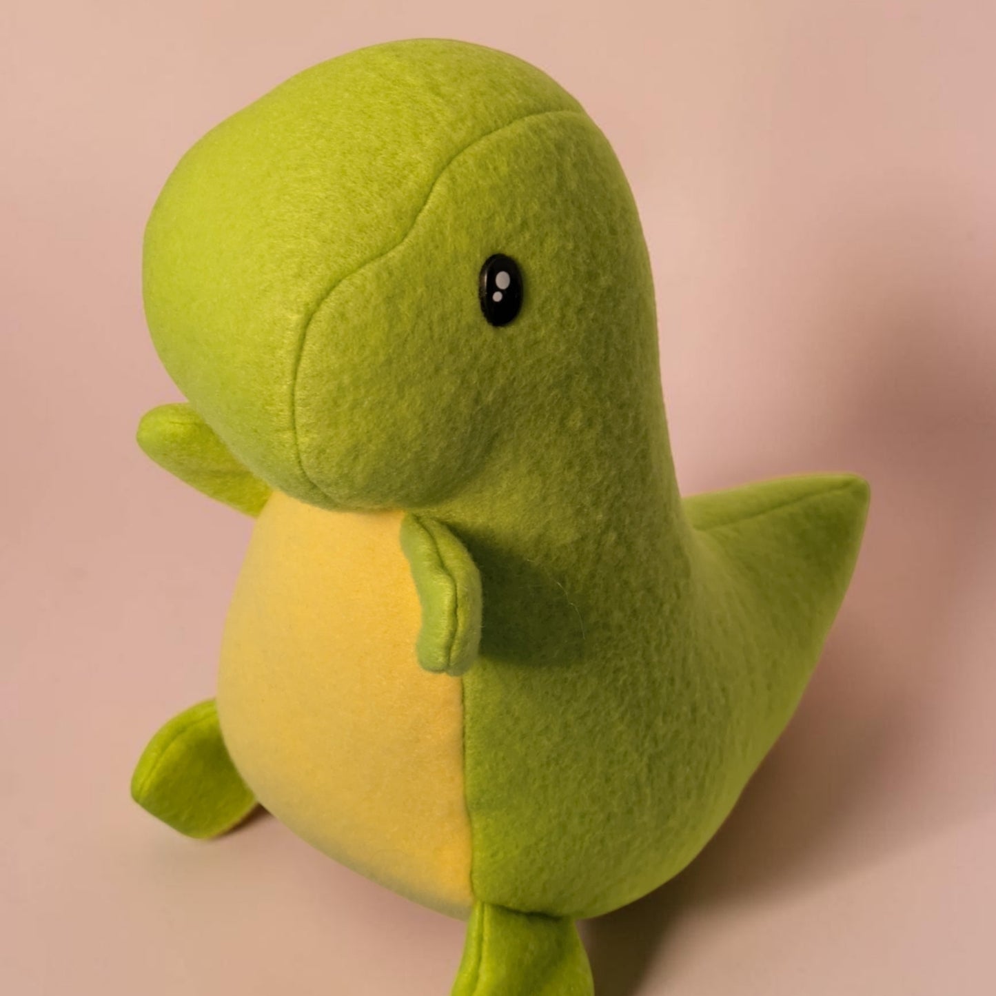 A small plush dinosaur toy in bright lime green with a pale yellow belly, photographed against a soft pink background.
