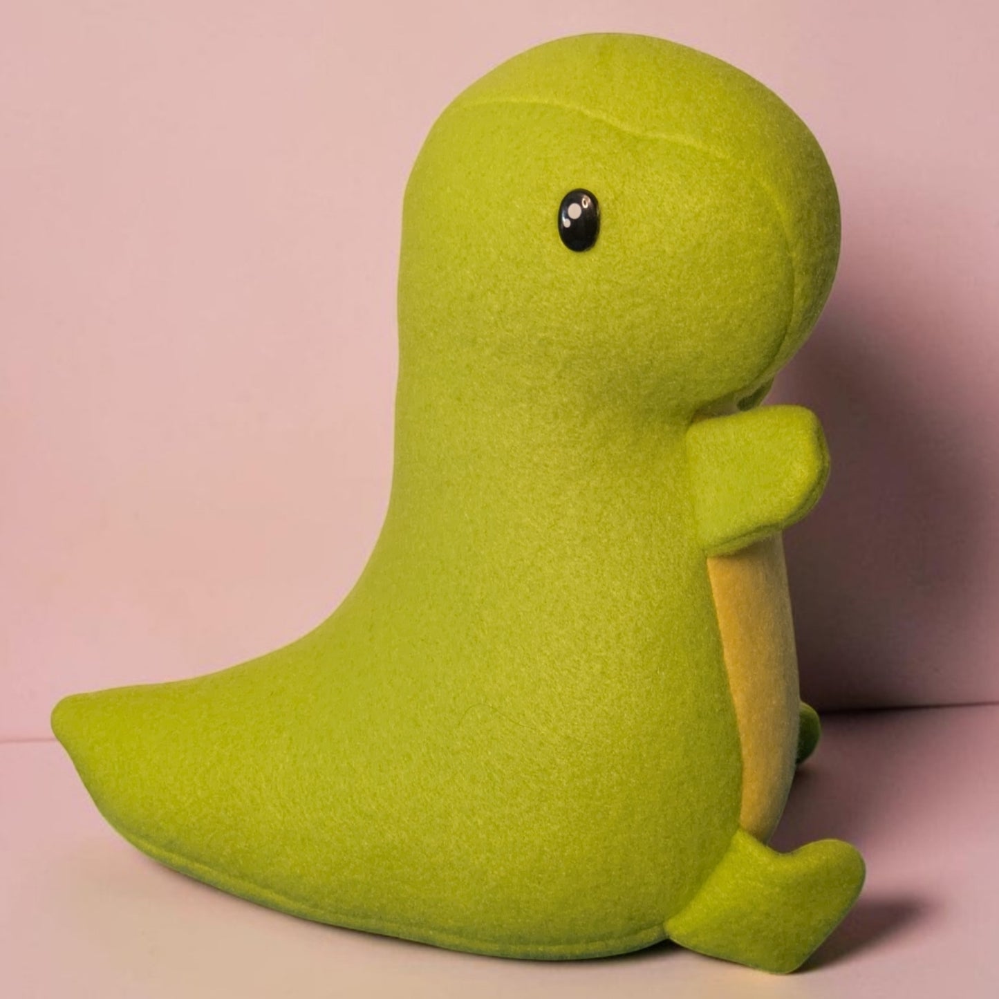 A cute lime green fleece dinosaur plush toy with a small black safety eye, sitting against a soft pink background.