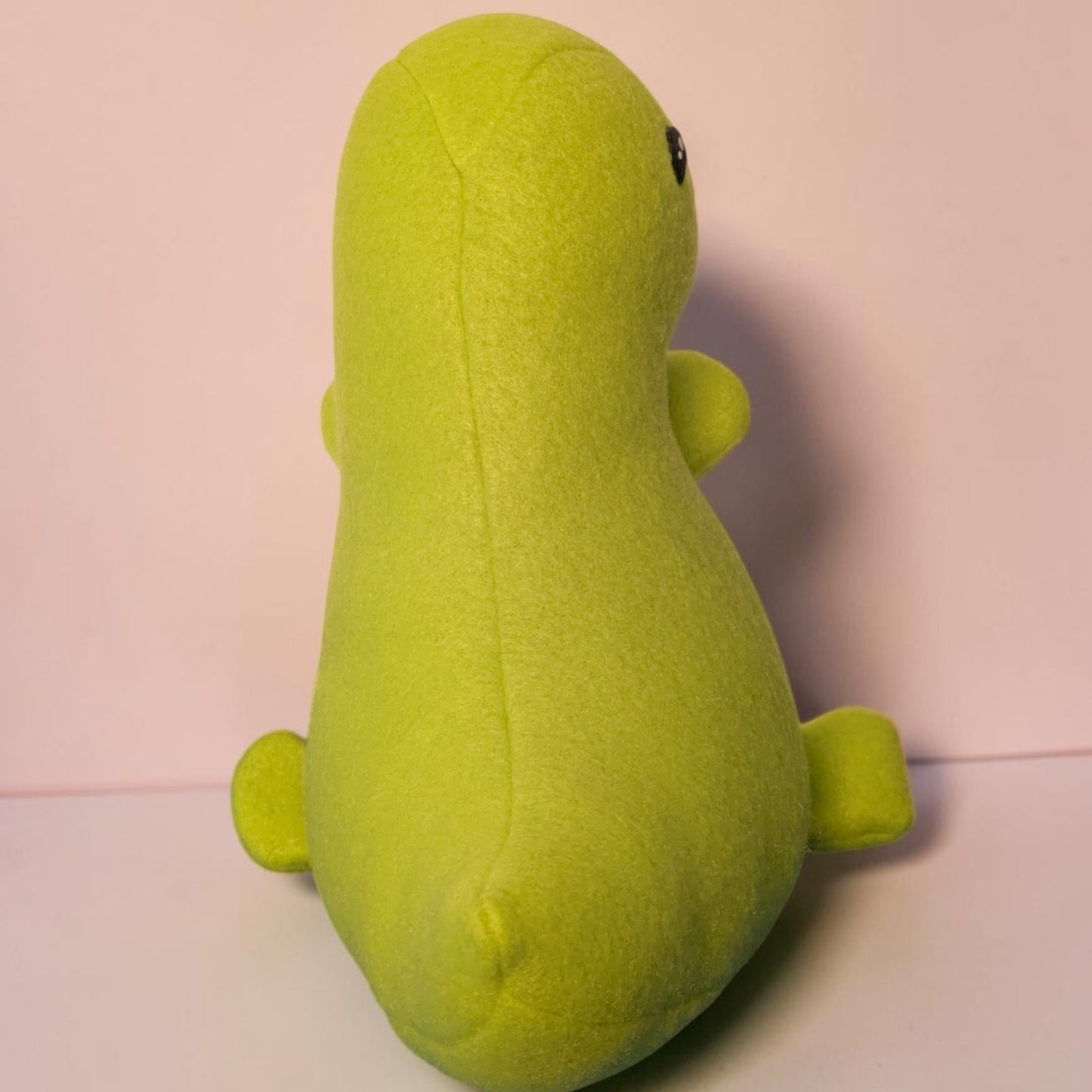 A lime green plush dinosaur toy shown from the back, featuring a soft fleece texture, small black eyes, and tiny limbs, set against a pale pink background.