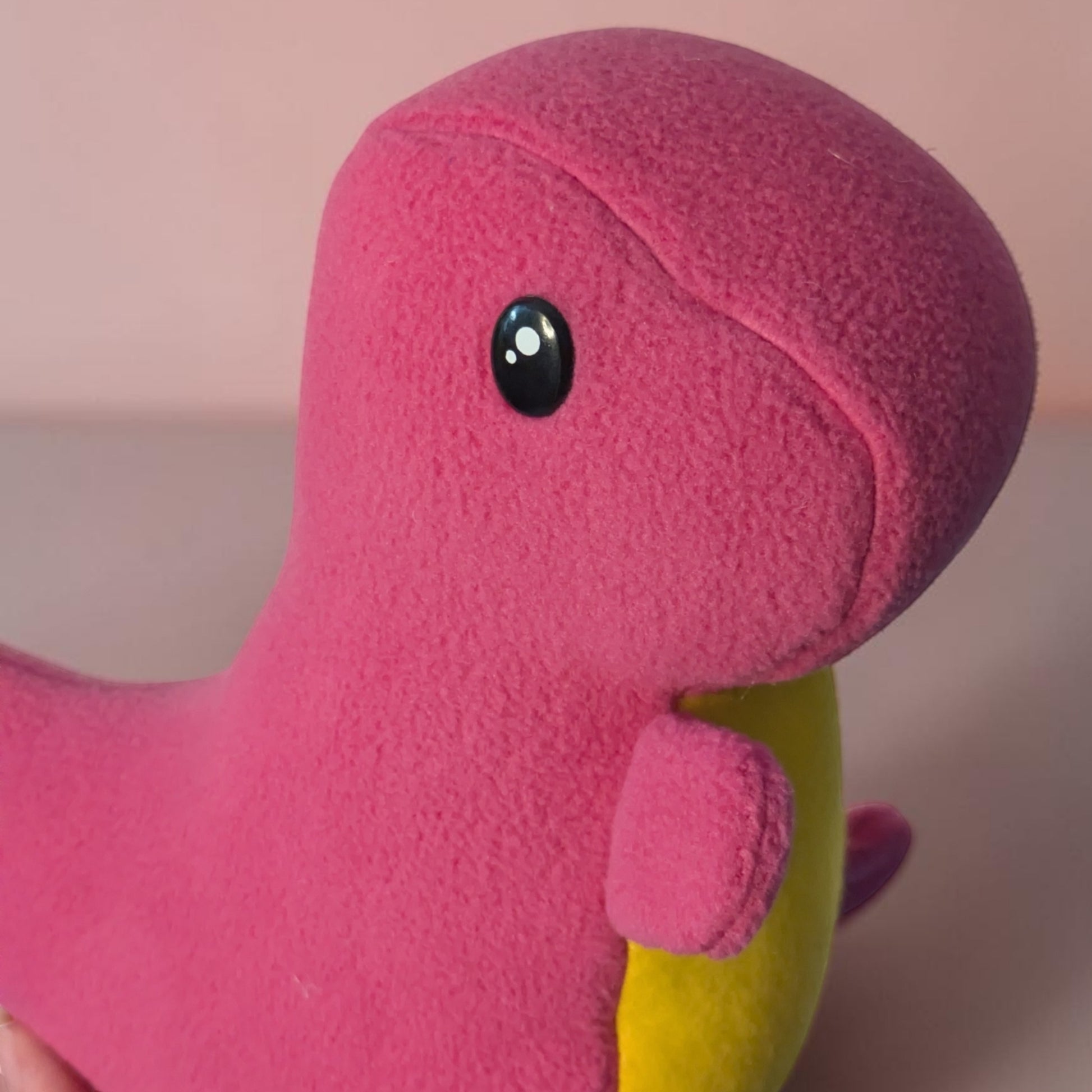 A close-up of a pink plush dinosaur toy with a yellow belly and a glossy black safety eye, shown against a soft pink background.
