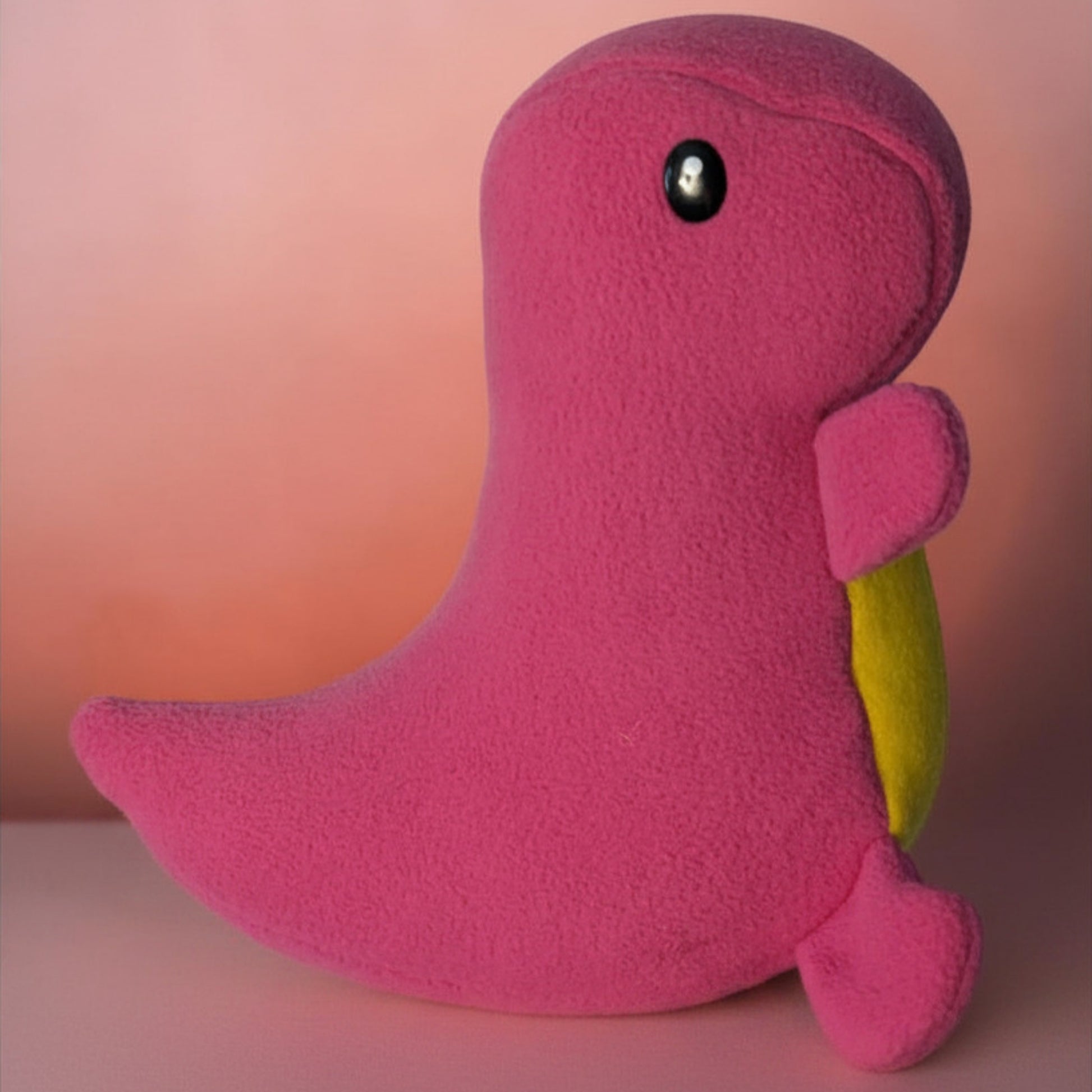 A pink plush dinosaur toy with a small yellow belly patch and a black safety eye, photographed against a soft pink gradient background.