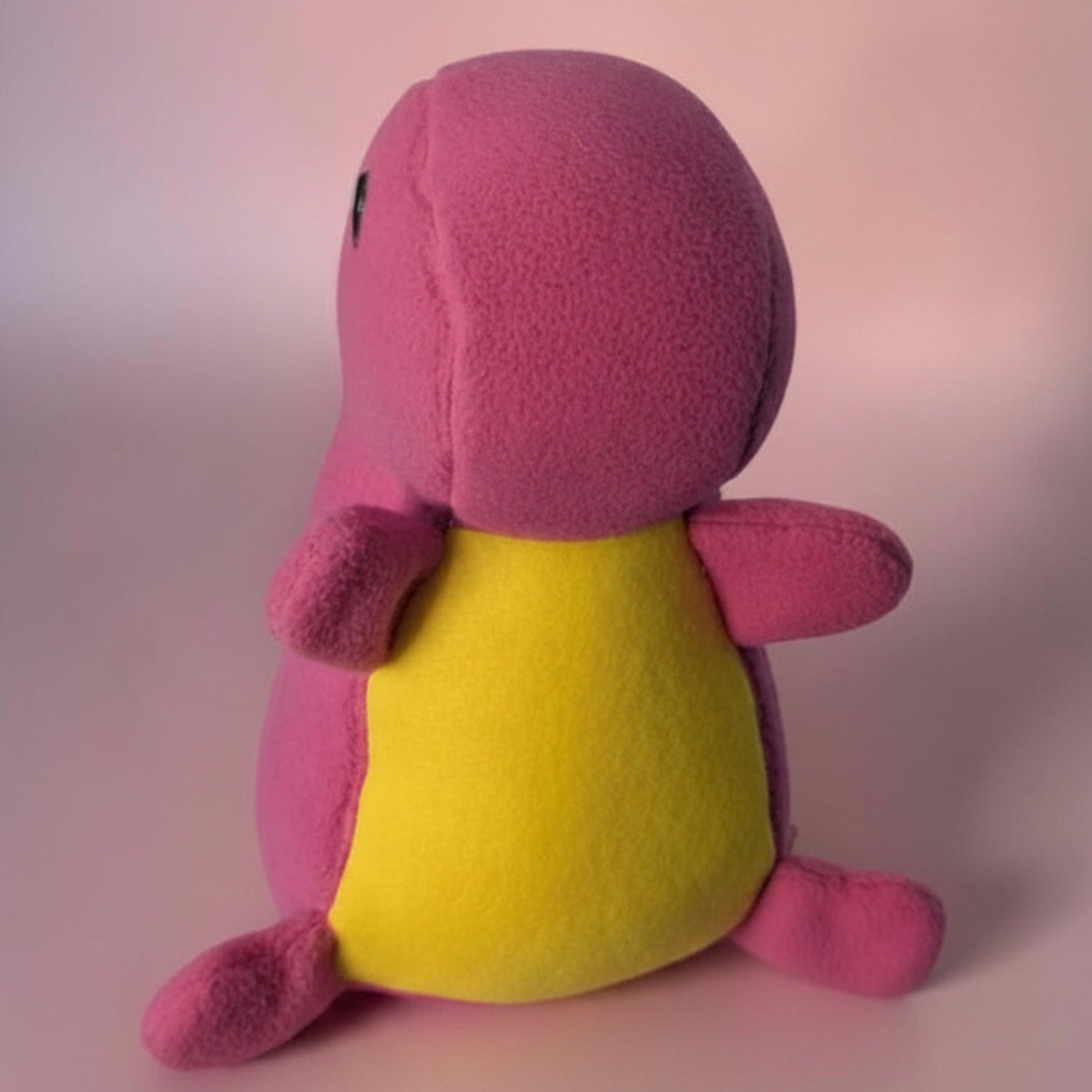 A pink and yellow plush dinosaur toy shown from the front, sitting against a soft pink studio background.
