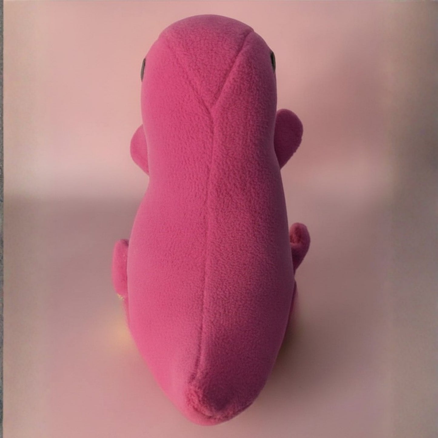 A pink plush toy shaped like a stylized dinosaur, photographed from the back against a soft pink background.