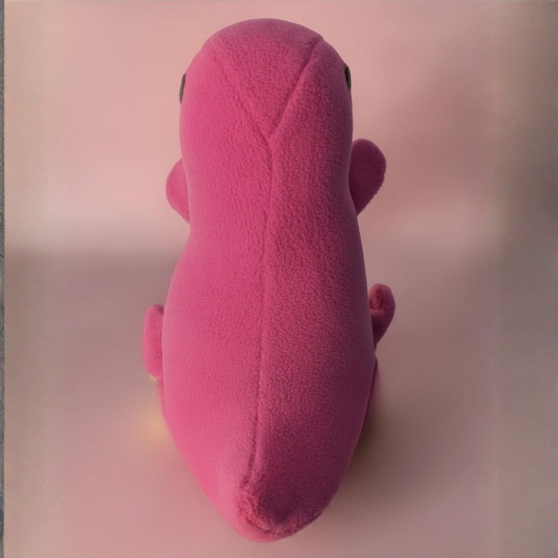 A pink plush toy shaped like a stylized dinosaur, photographed from the back against a soft pink background.