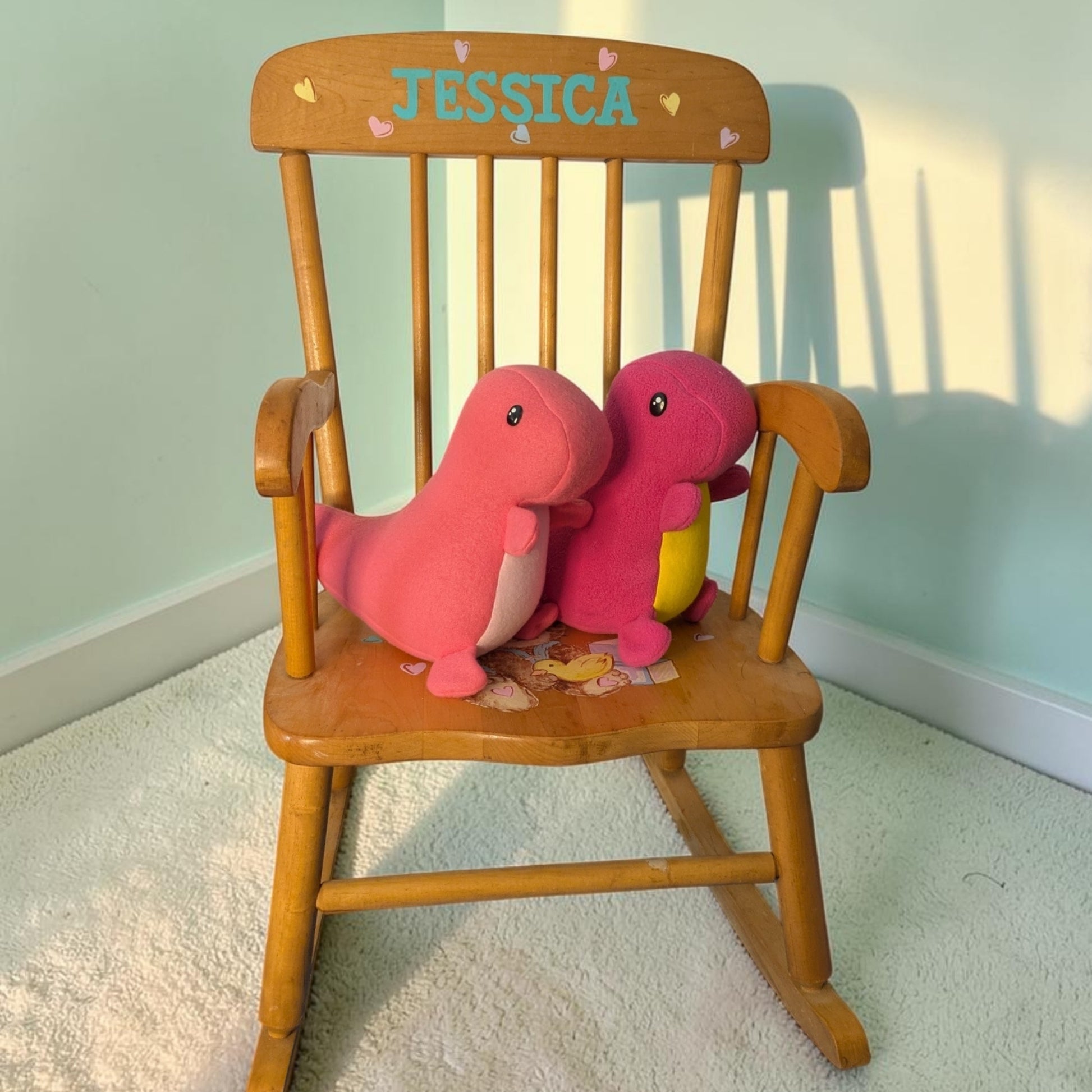 A wooden rocking chair with the name 'JESSICA' painted on the backrest, styled with two pink handmade plush dinosaur toys on a white carpeted floor against a pale green wall.