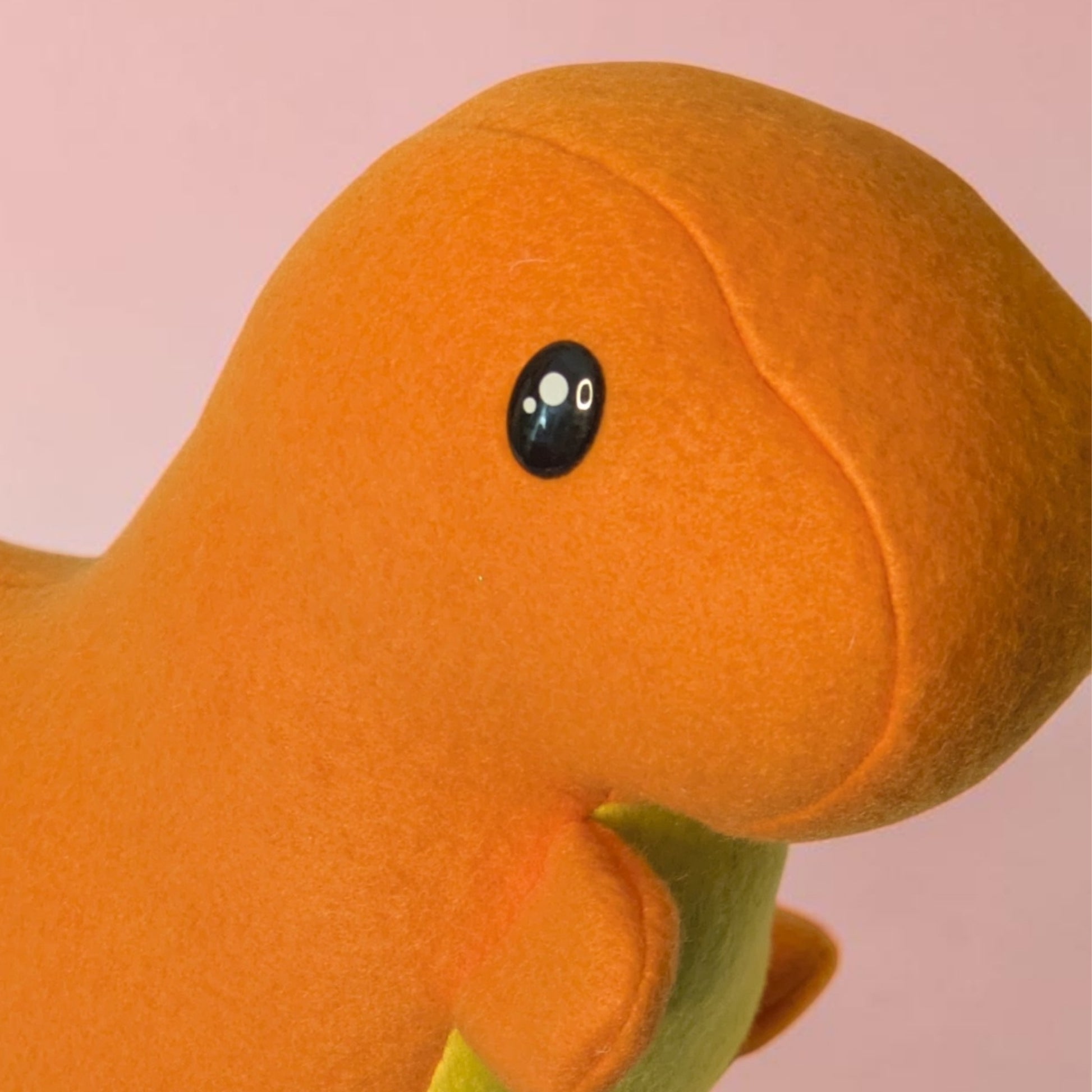 Close-up of an orange plush dinosaur toy with a black and white eye, set against a solid pink background.