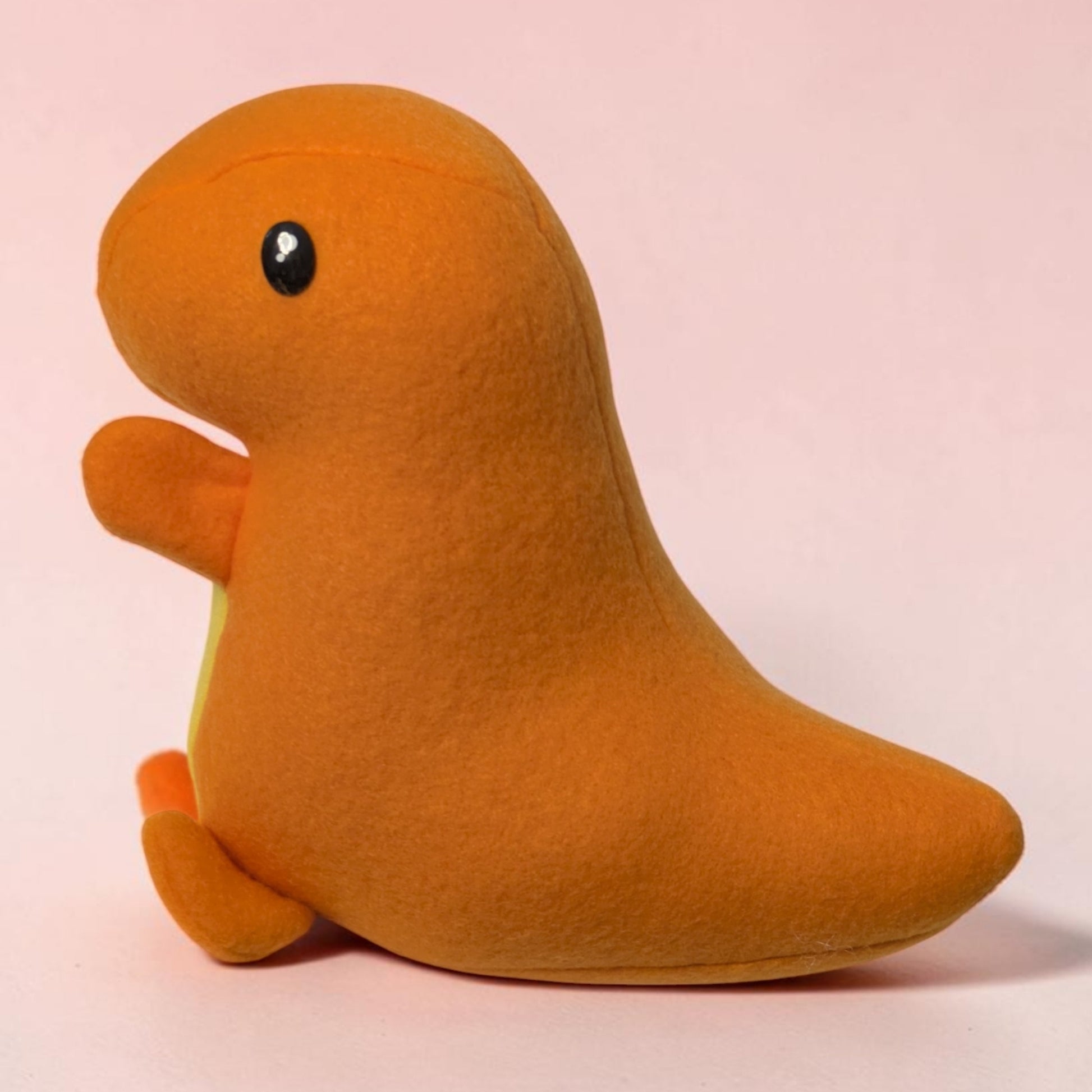 A handmade plush dinosaur  toy with an orange body, yellow belly and black eye, photographed against a soft light pink background.