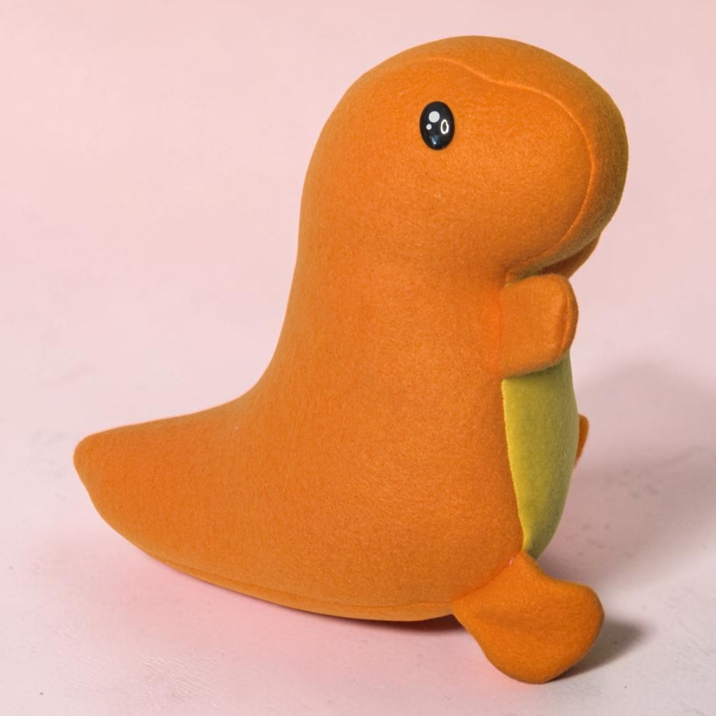 An orange dinosaur-shaped plush toy with a small yellow belly patch and black eyes, standing on a light pink background.