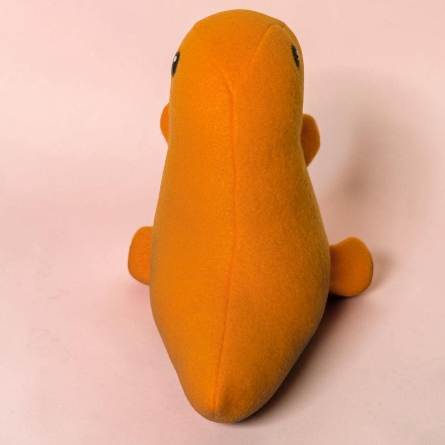 A small orange plush toy shaped like a dinosaur, photographed from the back on a plain light pink background.
