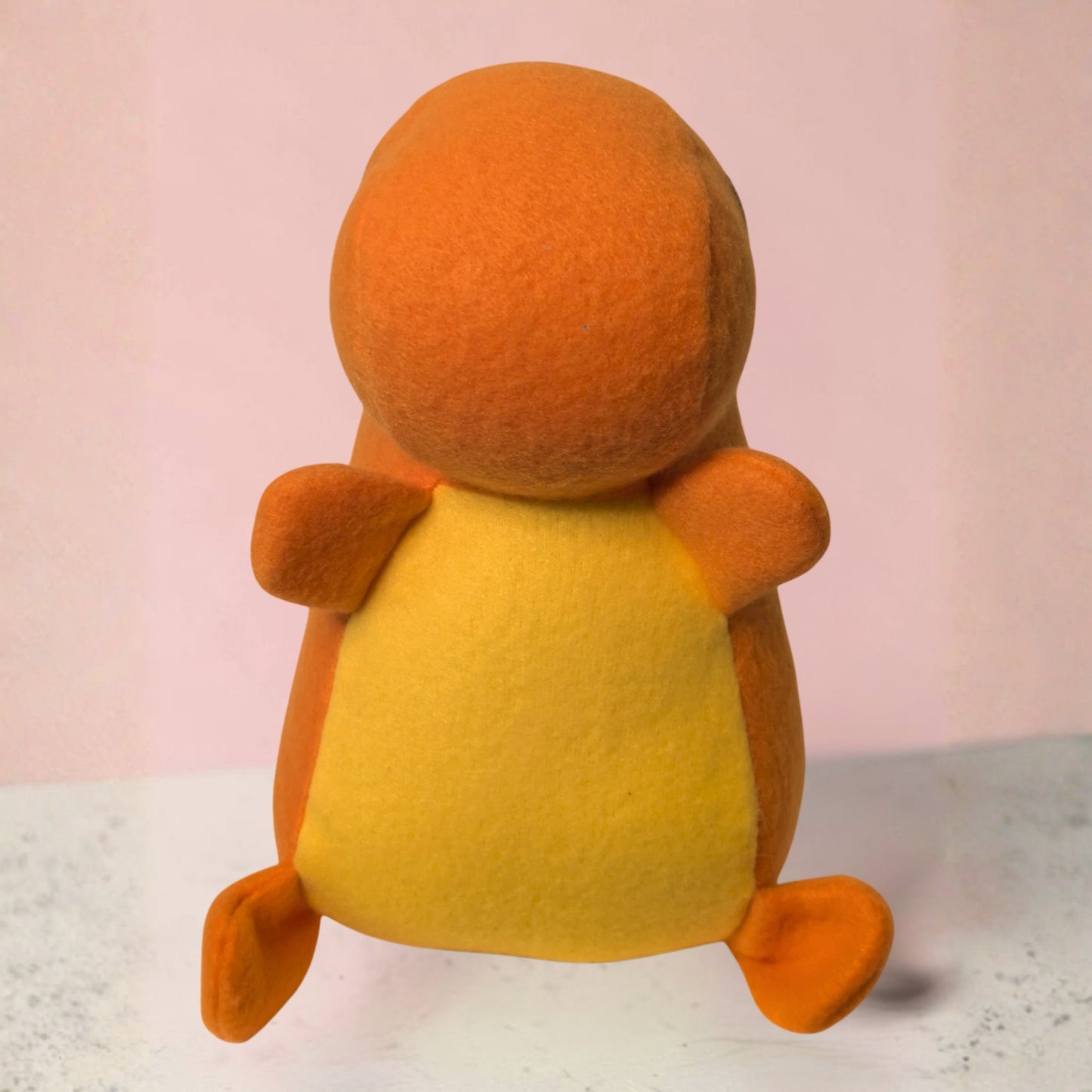 A fleece plush toy shaped like a small orange and yellow dinosaur, photographed from the front against a soft pink background.