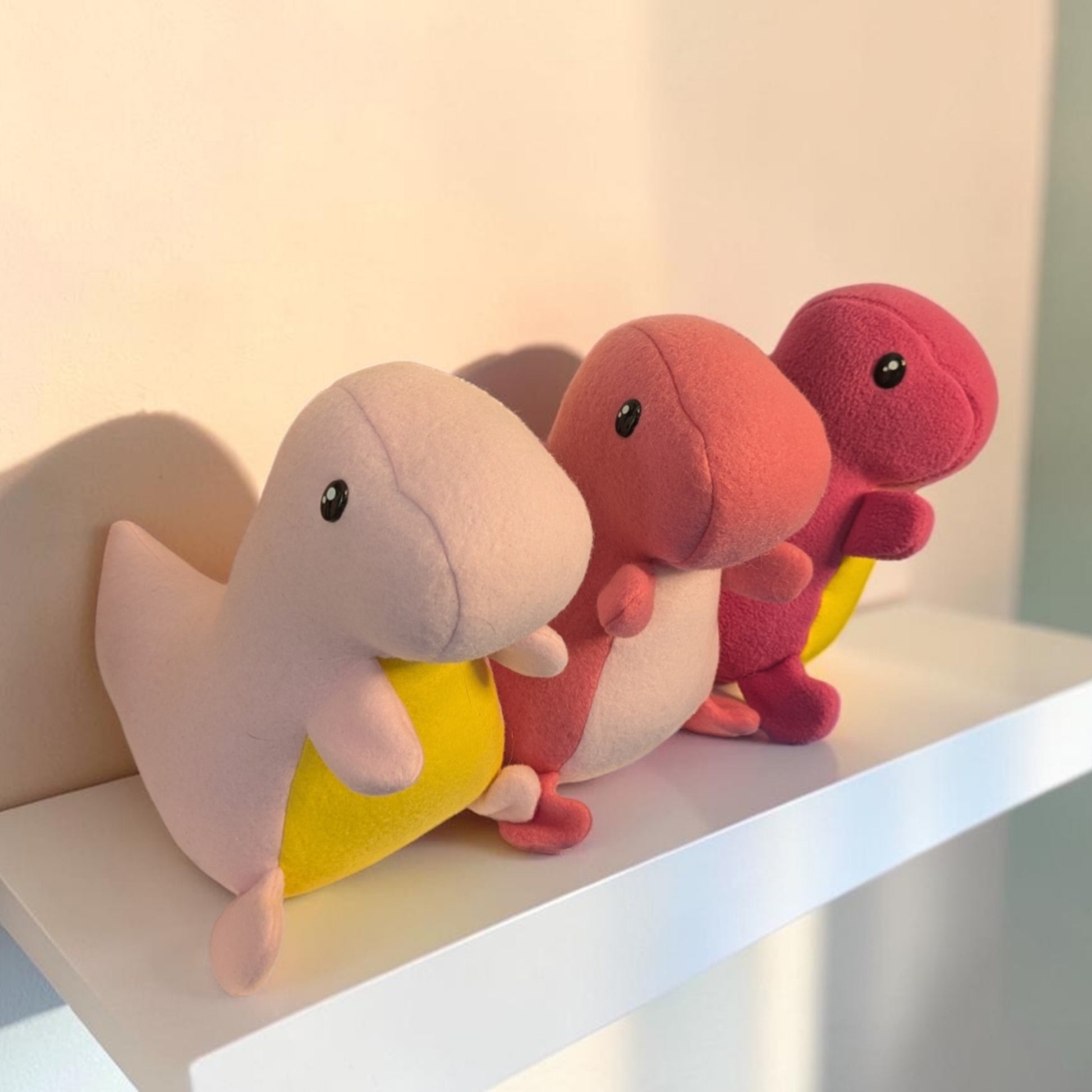 Three small plush dinosaur toys in pastel pink, coral pink, and magenta, each has a yellow belly, arranged on a white shelf against a softly lit beige wall.