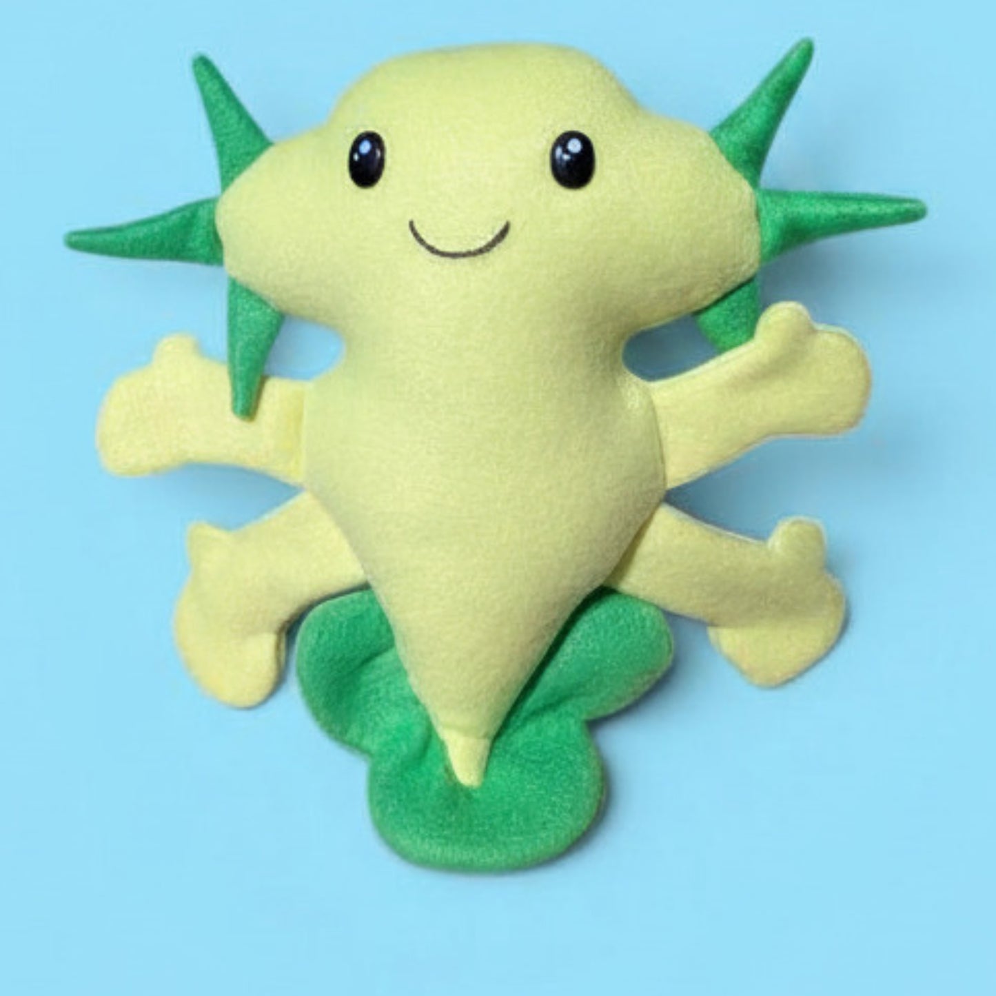 A handmade plush toy of a cute, smiling light green axolotl with light green body and darker green fins and gills, set against a solid light blue background.