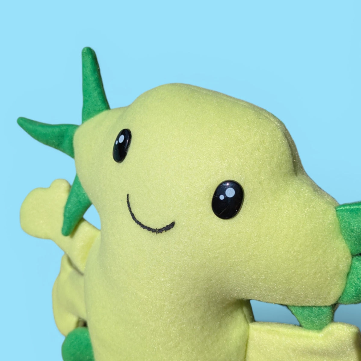 A close up of a green axolotl plush stuffed animals face with black safety eyes and a smile.