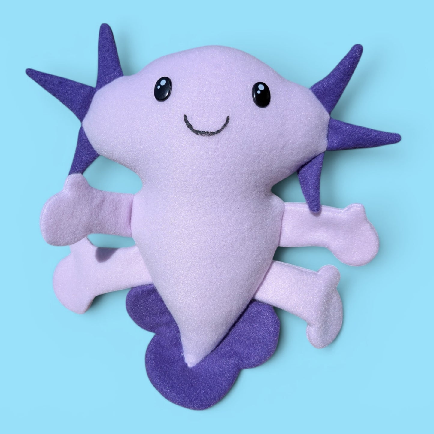 A cute, soft plush toy shaped like a smiling axolotl, featuring a light lavender body with dark purple fins and gills, set against a solid light blue background.