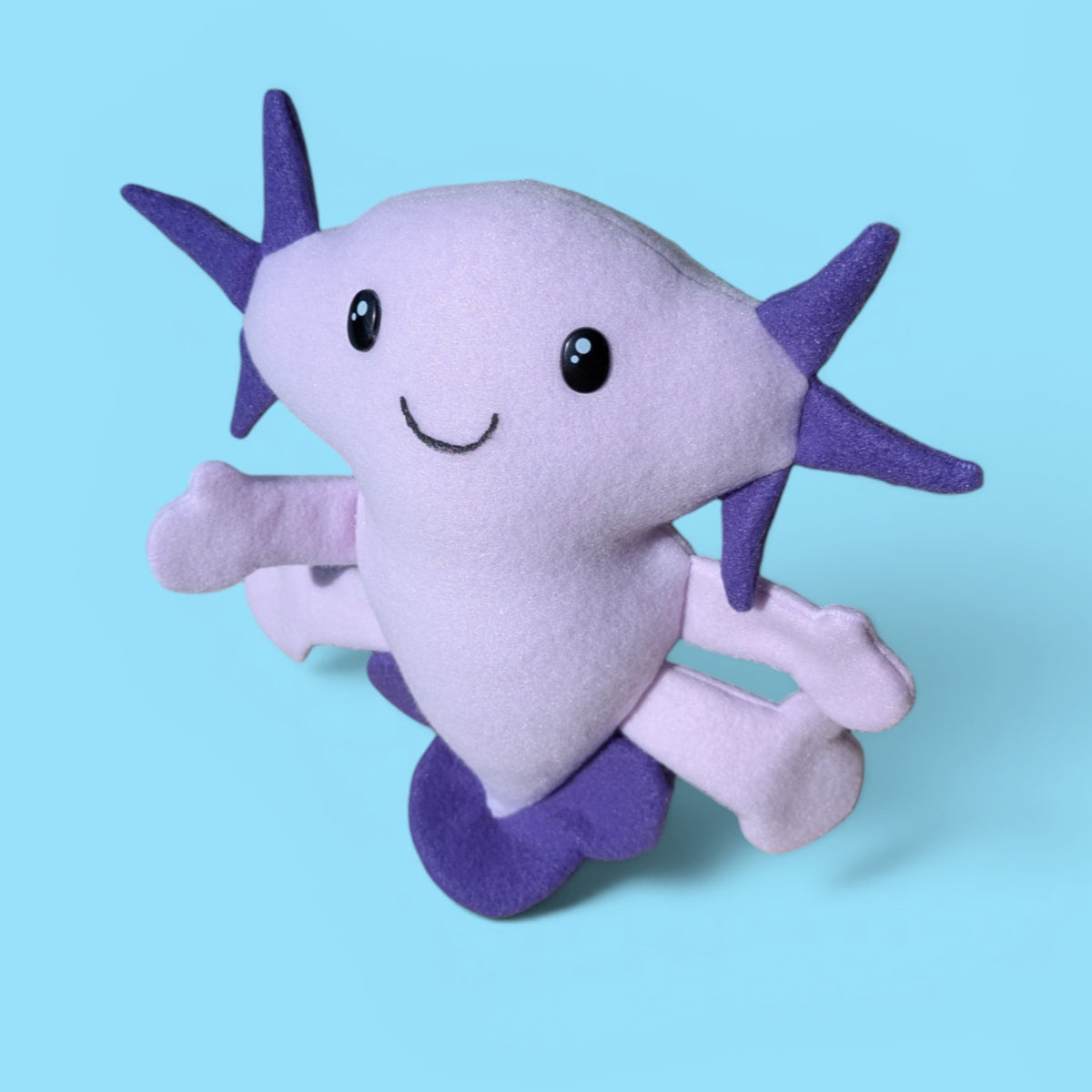 A plush toy shaped like a smiling axolotl, made of soft lavender and purple felt, set against a solid light blue background.