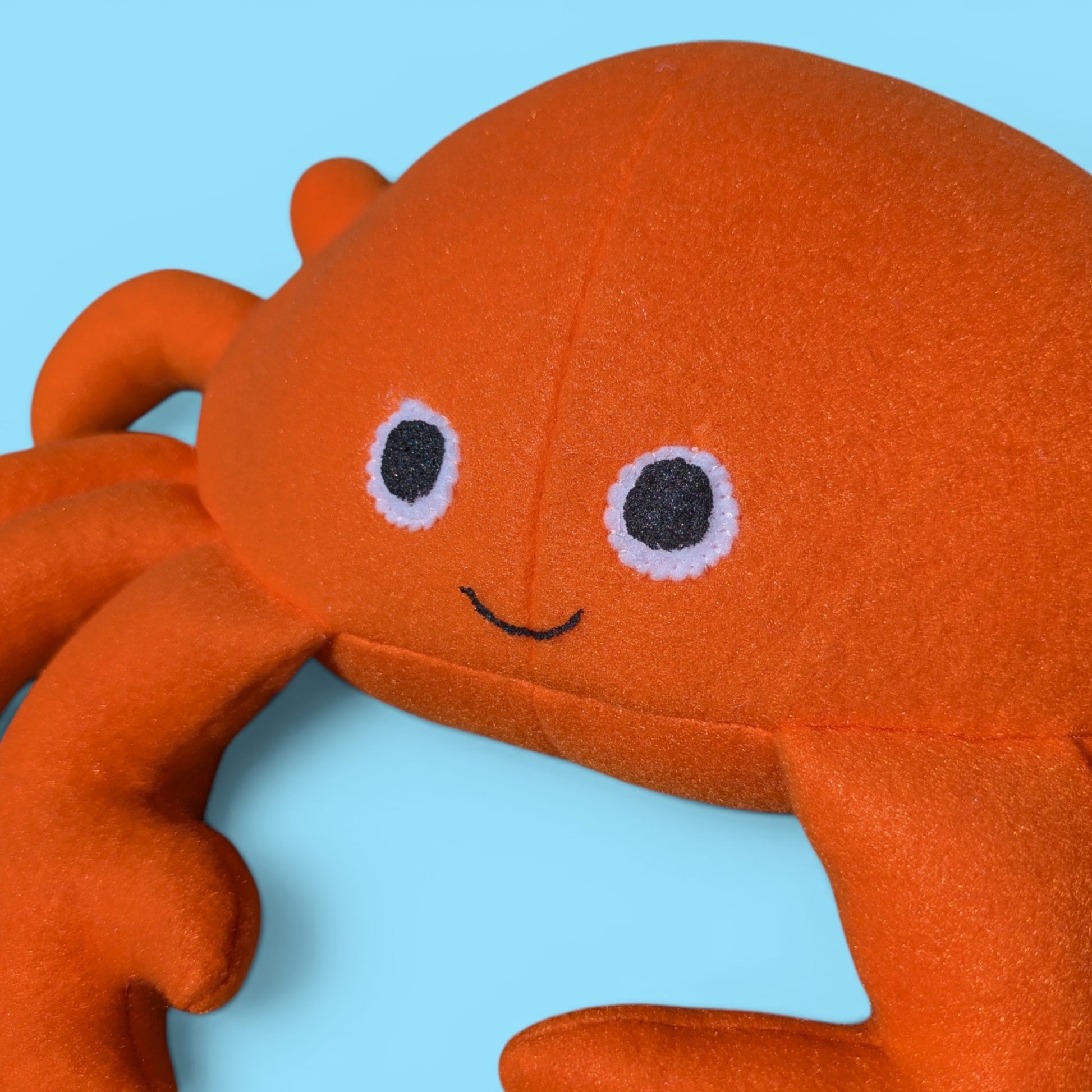 A close-up of an orange plush crab toy with felt eyes and a smile, set against a light blue background.