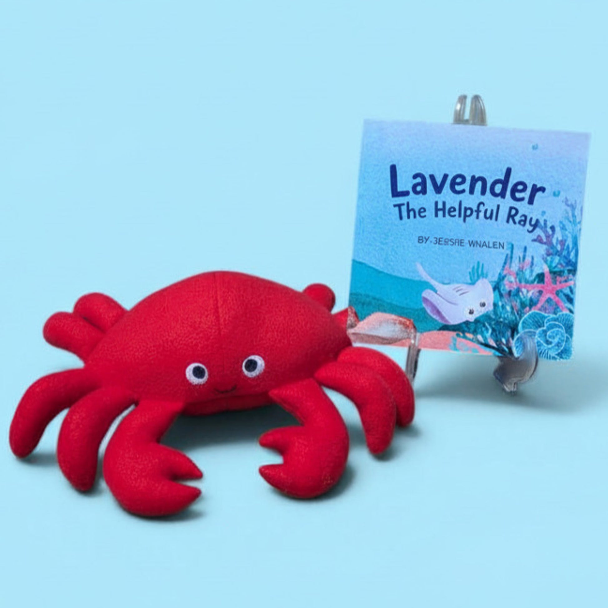 A red plush crab toy sits in front of a small book titled "Lavender The Helpful Ray" by Jersie Mwalen, displayed on a clear stand against a light blue background.