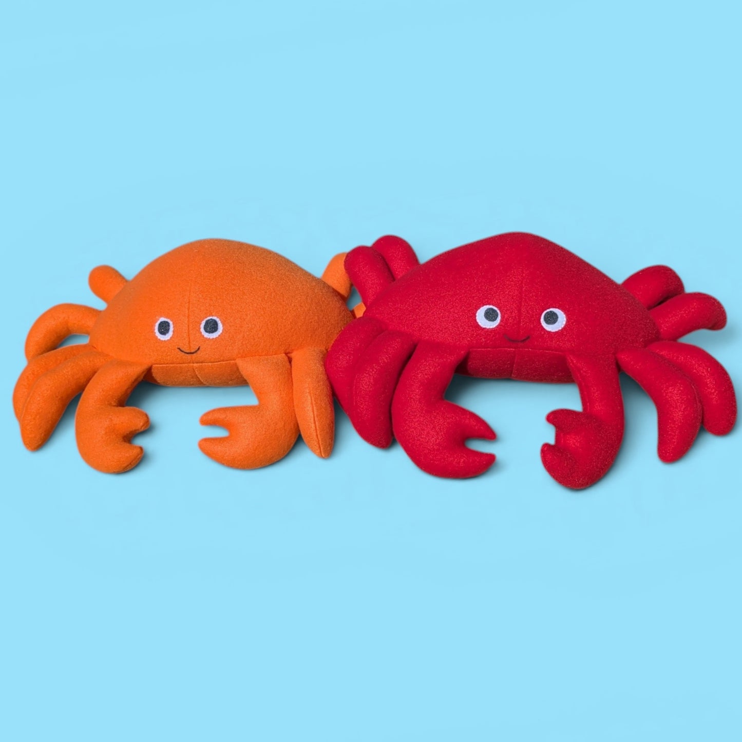 Two plush crab toys side by side on a solid light blue background. The left crab is orange, and the right crab is red, both with felt eyes and simple facial features.