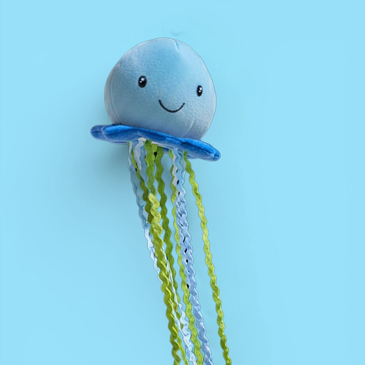 A cute jellyfish wall hook featuring a light blue plush head with a smiling face, a blue ring collar, and wavy green, yellow, and blue hanging tentacles, shown against a solid light blue background.