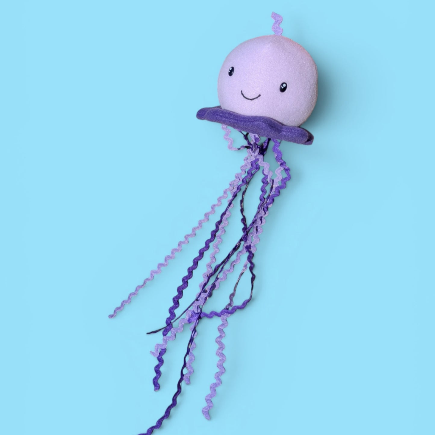 A plush jellyfish toy with a light purple round head, a darker purple bell, and long wavy purple tentacles, shown against a solid light blue background.