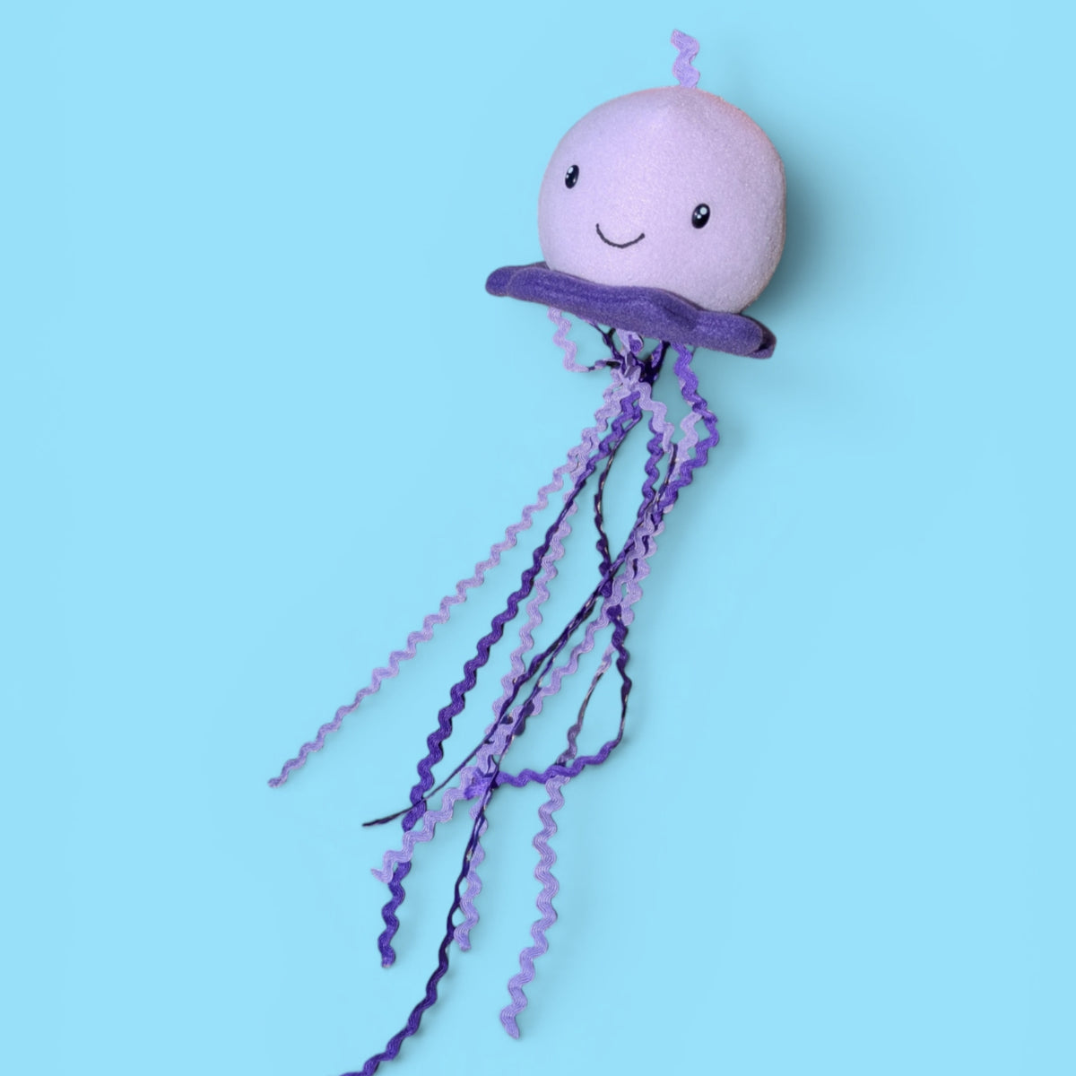 A plush jellyfish toy with a light purple round head, a darker purple bell, and long wavy purple tentacles, shown against a solid light blue background.