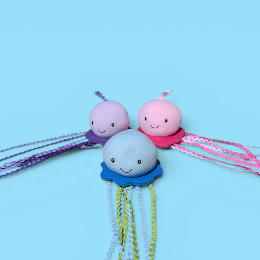 Three small jellyfish-shaped plush toys arranged together on a light blue background. The front jellyfish is light blue with a blue bell and yellow-green tentacles, while the two behind it are pink and lavender with purple and pink tentacles.
