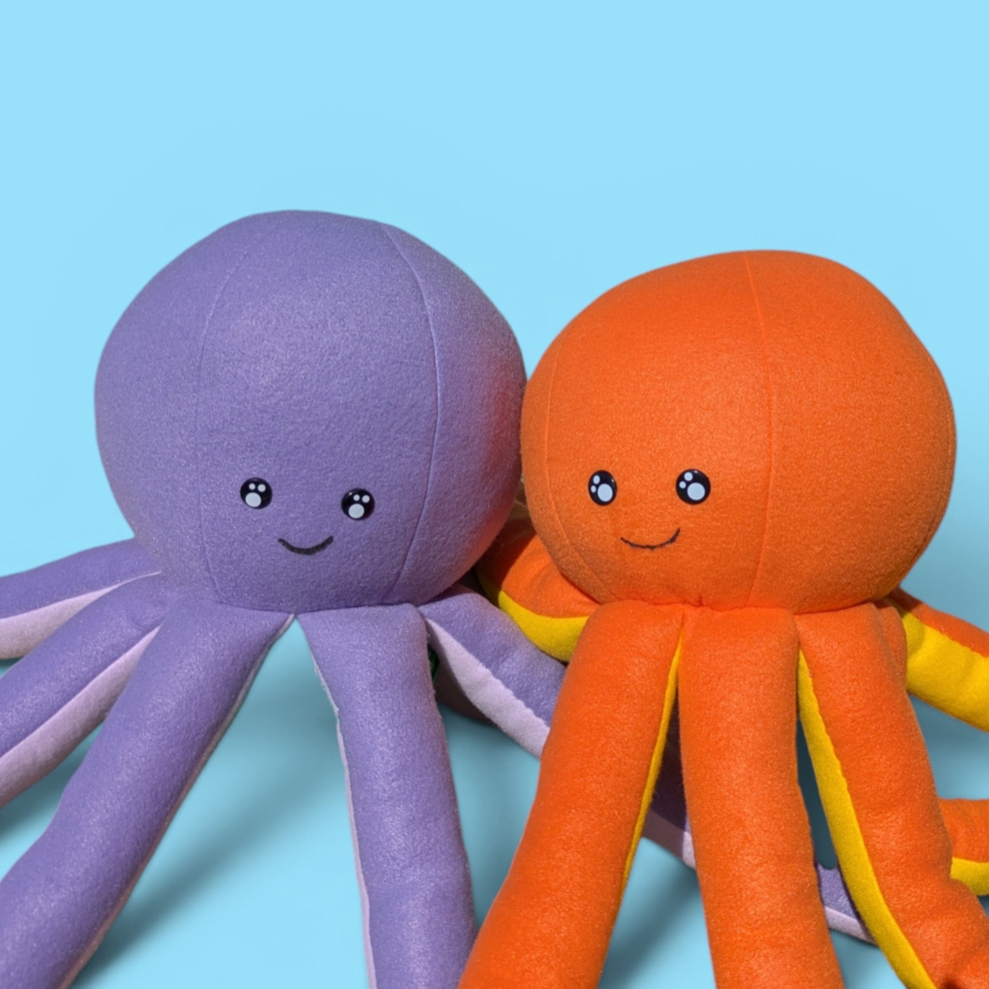 Two plush octopus toys made of soft fabric, one purple and one orange, both with smiling faces, set against a solid light blue background.