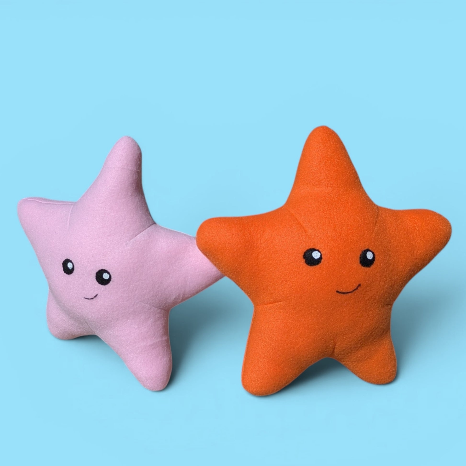 Two plush star-shaped stuffed animals on a light blue background, one in pink and one in orange, both with smiling faces.