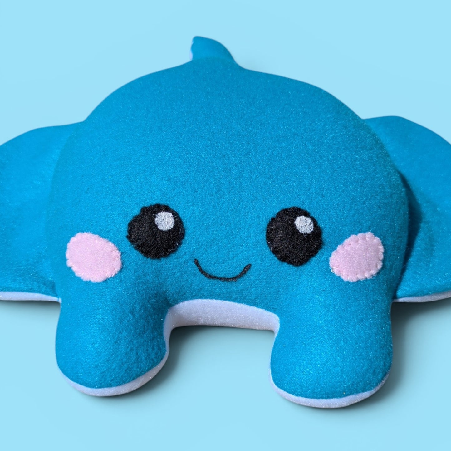 A blue felt sea creature plush stuffed animal with a smiling face, pink cheeks, and black eyes, set against a solid light blue background.