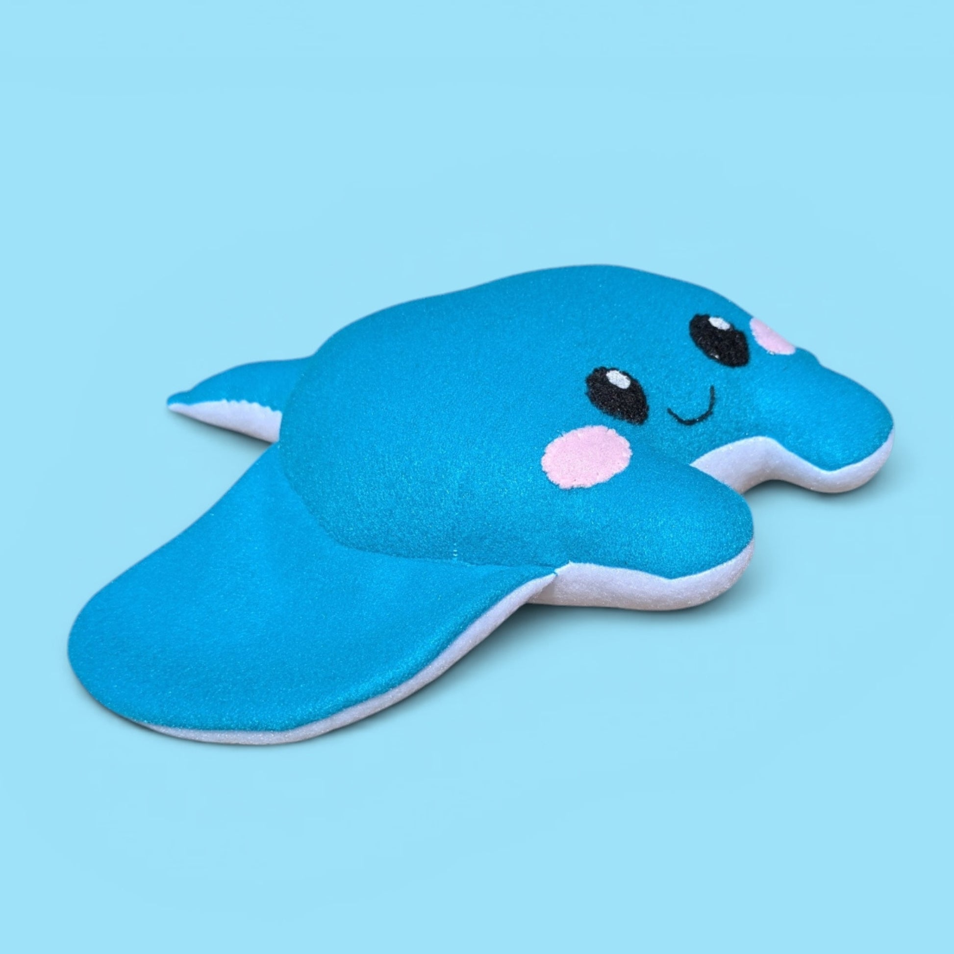 A blue plush stingray stuffed animal with embroidered black eyes, a smiling mouth, pink cheeks, and white belly, shown on a solid light blue background.