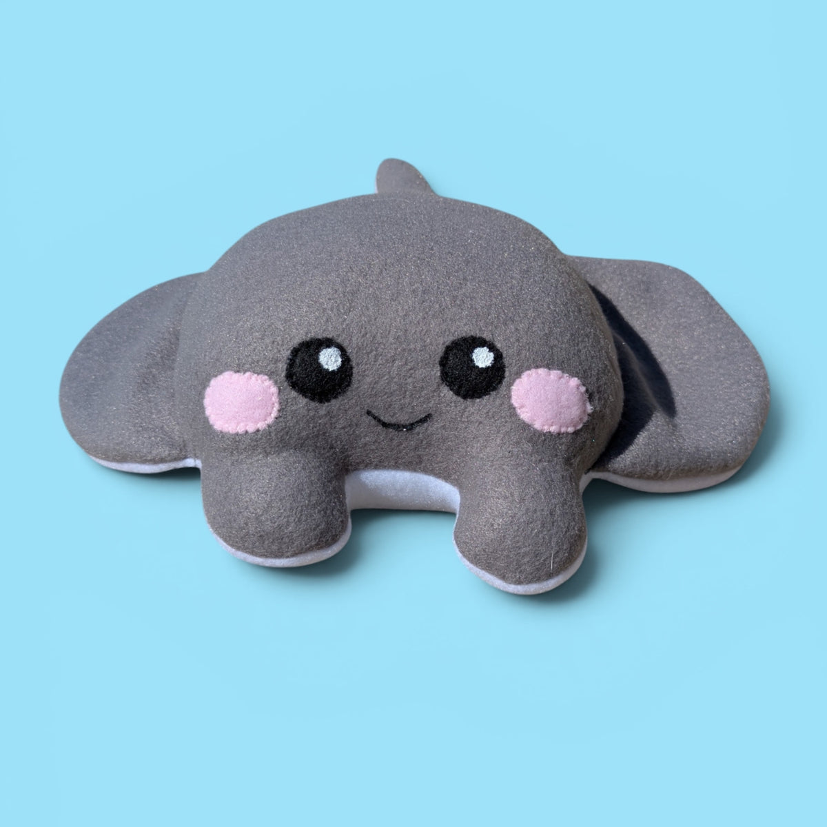 A plush stingray-shaped toy in dark gray with felt black eyes, pink cheeks, and a small smile, shown on a solid light blue background.