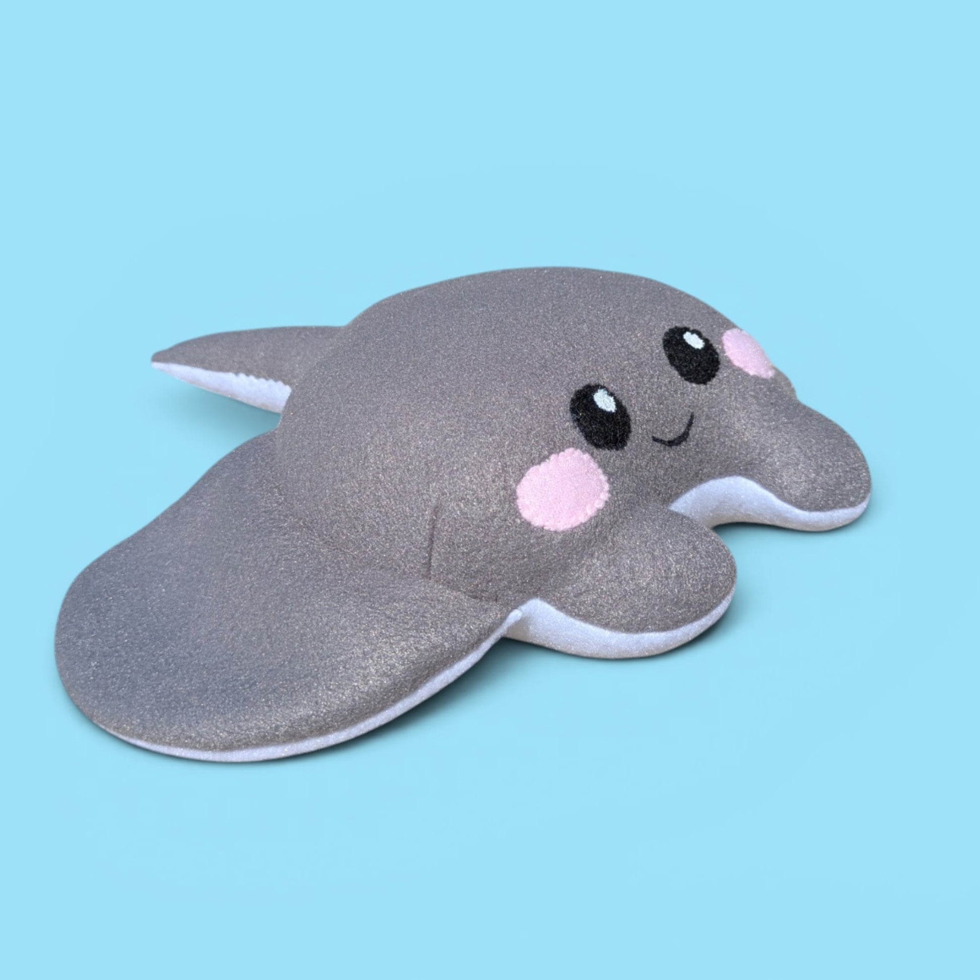 Side view of a plush gray stingray toy with a white underside and black eyes and pink cheeks