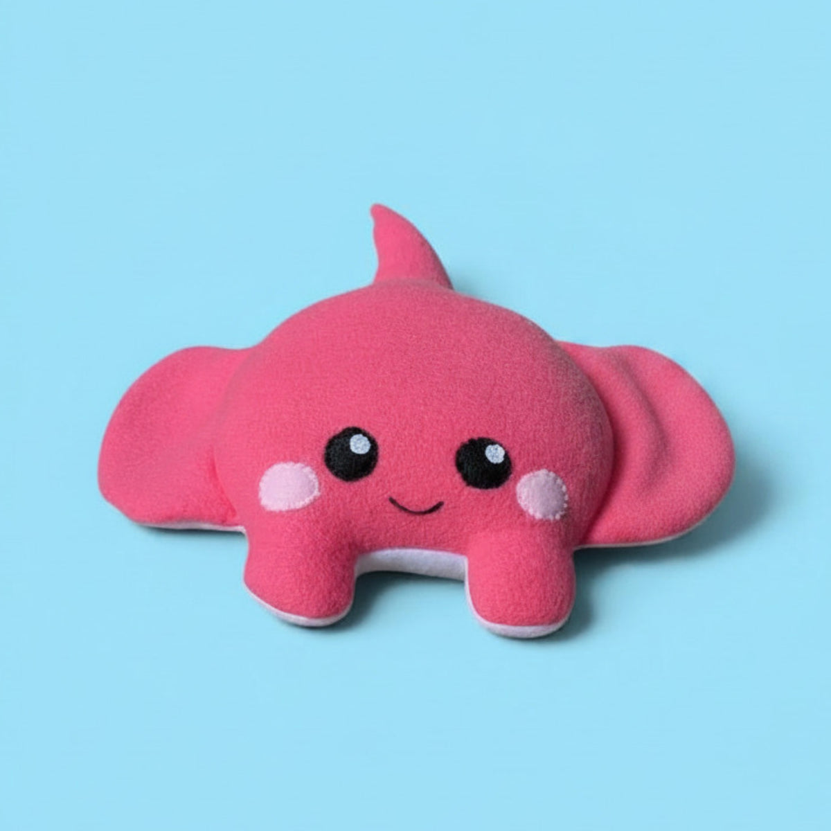 A pink plush stingray toy with large floppy wings, black felt eyes, a small smile, and pink cheek patches, shown on a light blue background.