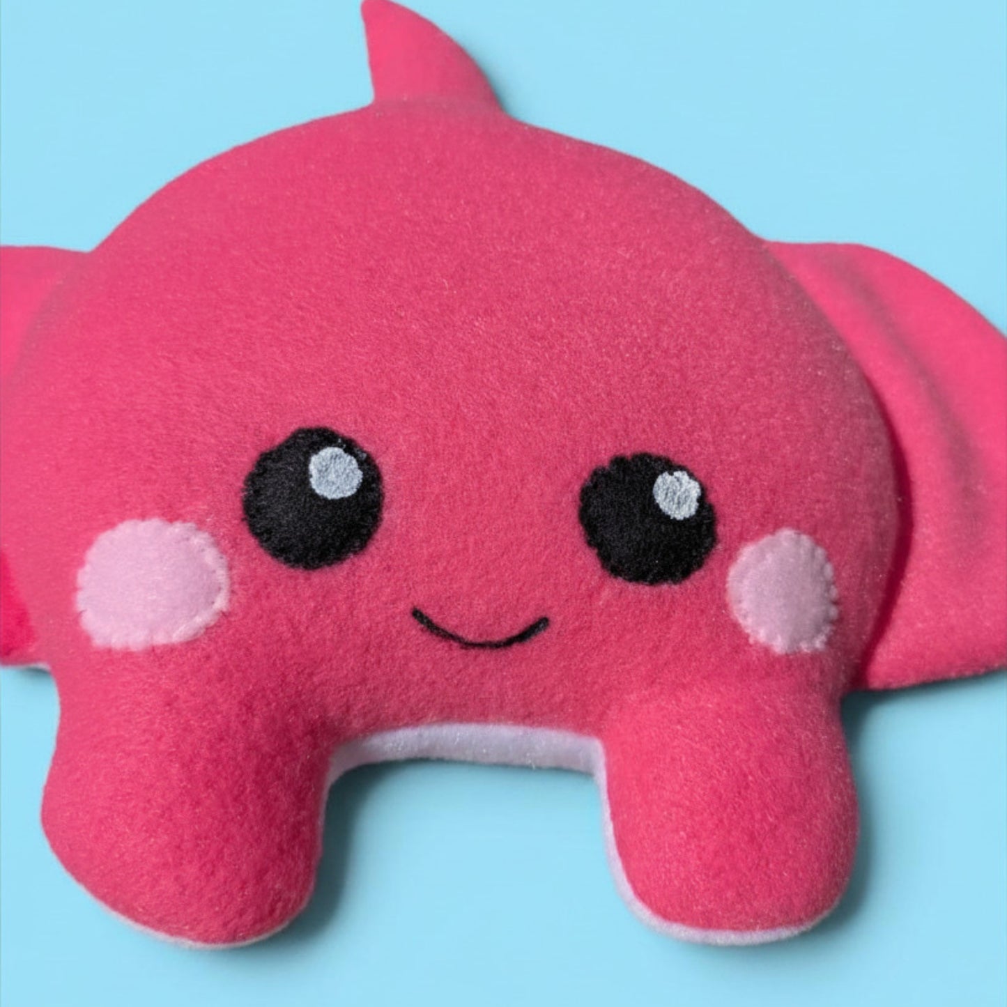 A pink plush stingray stuffed animal with embroidered eyes, a smile, and pink cheek patches, shown against a light blue background.
