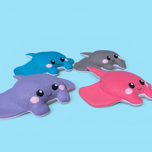 Four cute, soft plush stingray toys in purple, blue, gray, and pink, arranged on a solid light blue background.