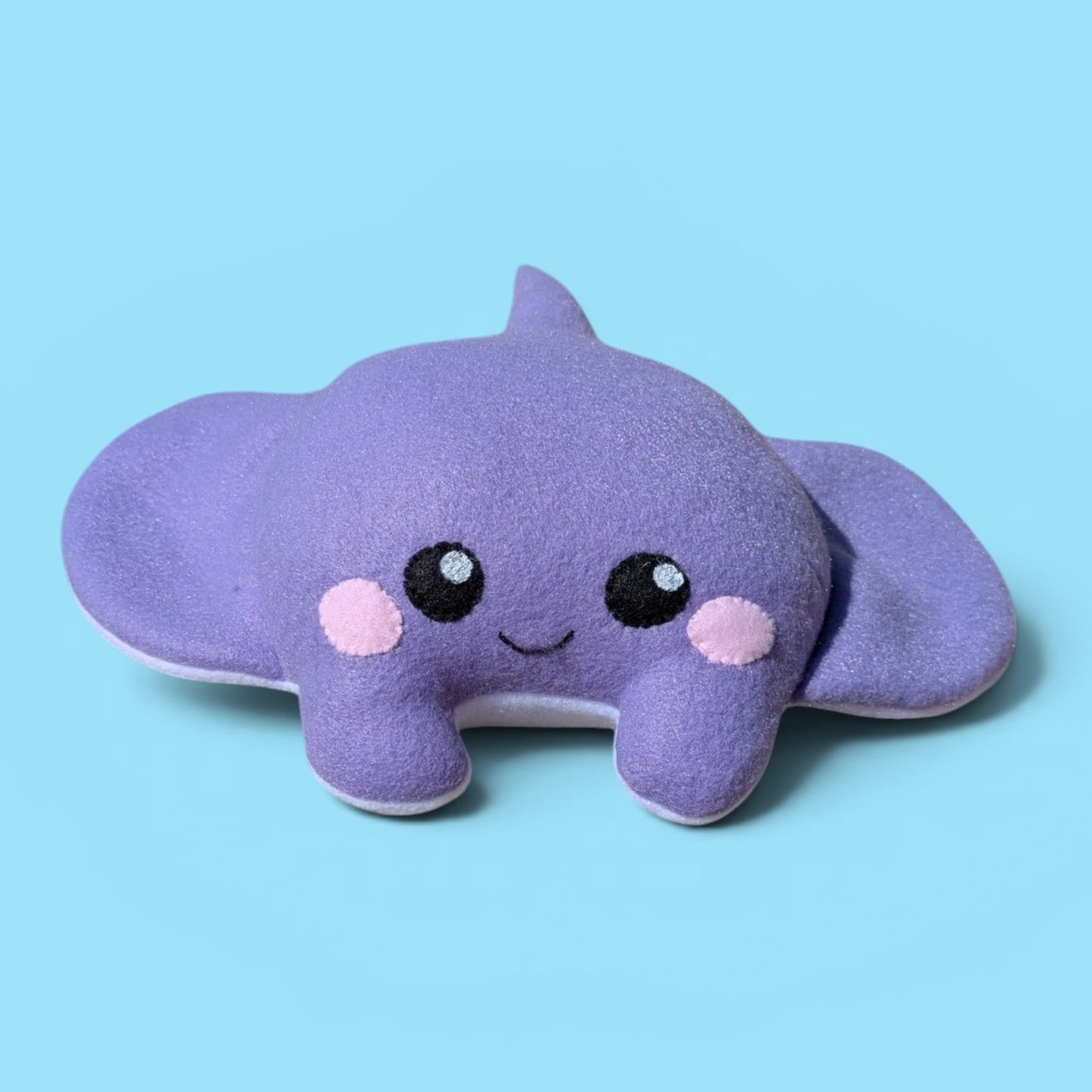 A cute purple plush stingray stuffed animal with felt eyes, a smiling mouth, pink cheeks, and a small tail, set against a solid light blue background.