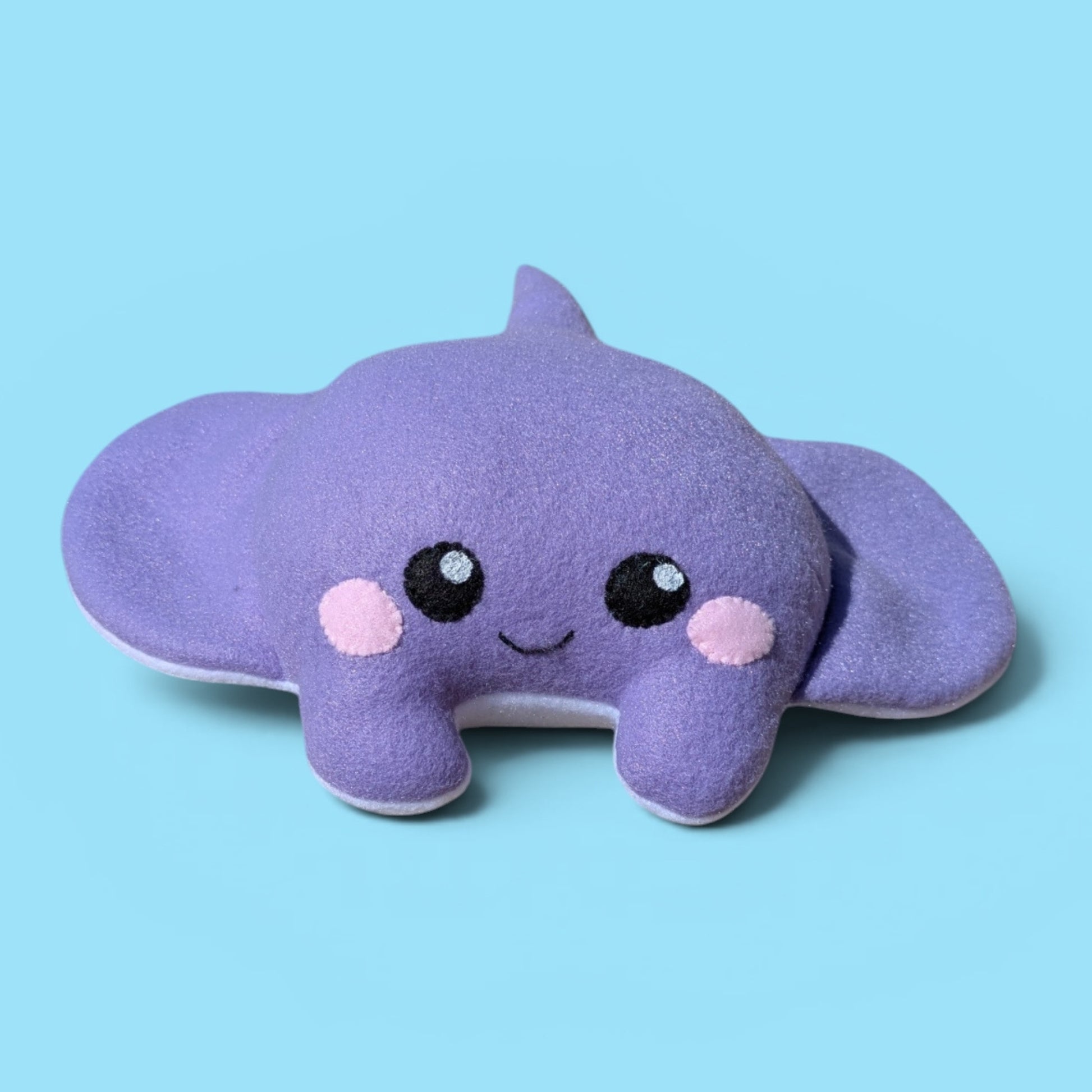 A cute purple plush stingray stuffed animal with felt eyes, a smiling mouth, pink cheeks, and a small tail, set against a solid light blue background.