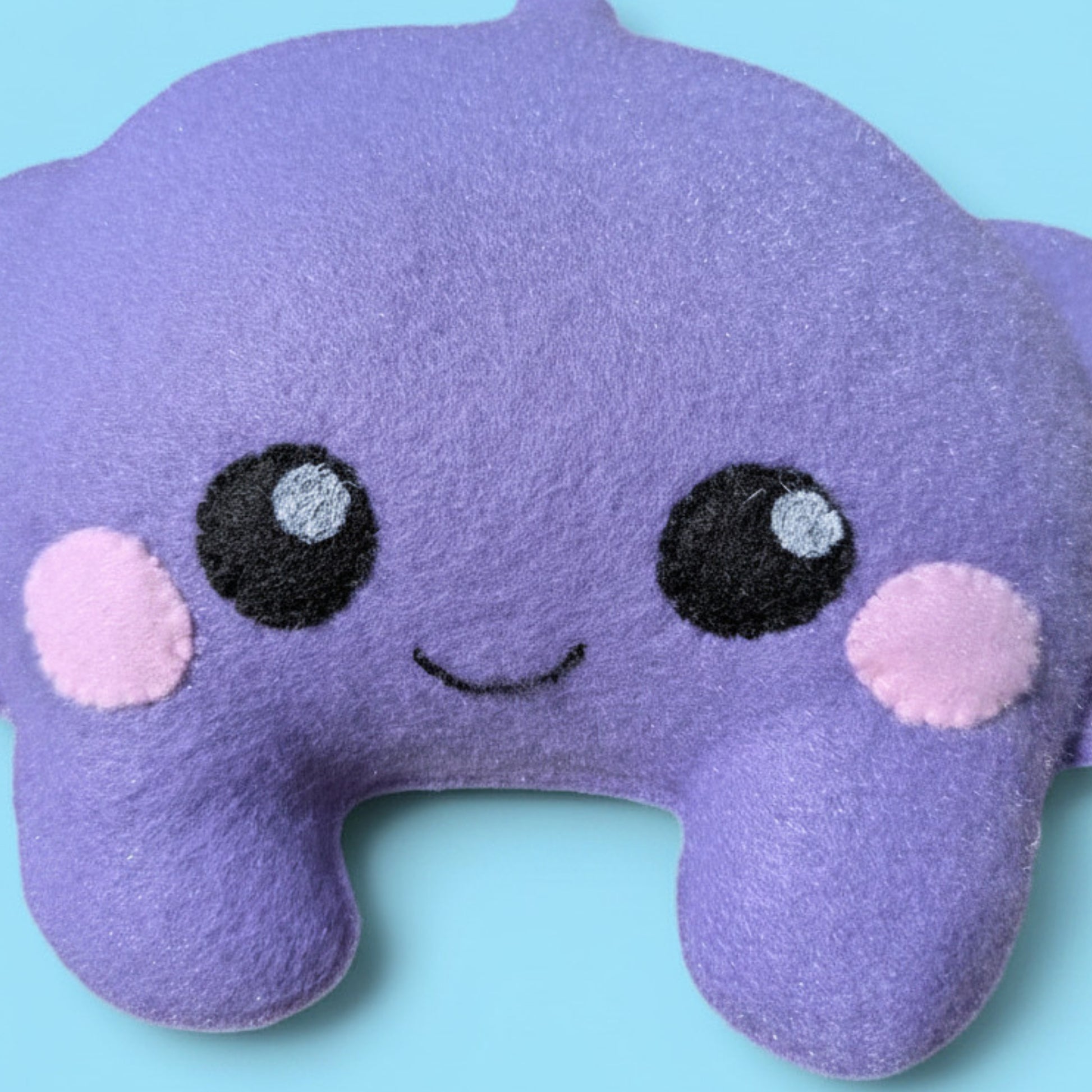 Close up picture of a fleece plush purple stingray stuffed animal. It has black felt eyes and pink cheeks and a gentle smile