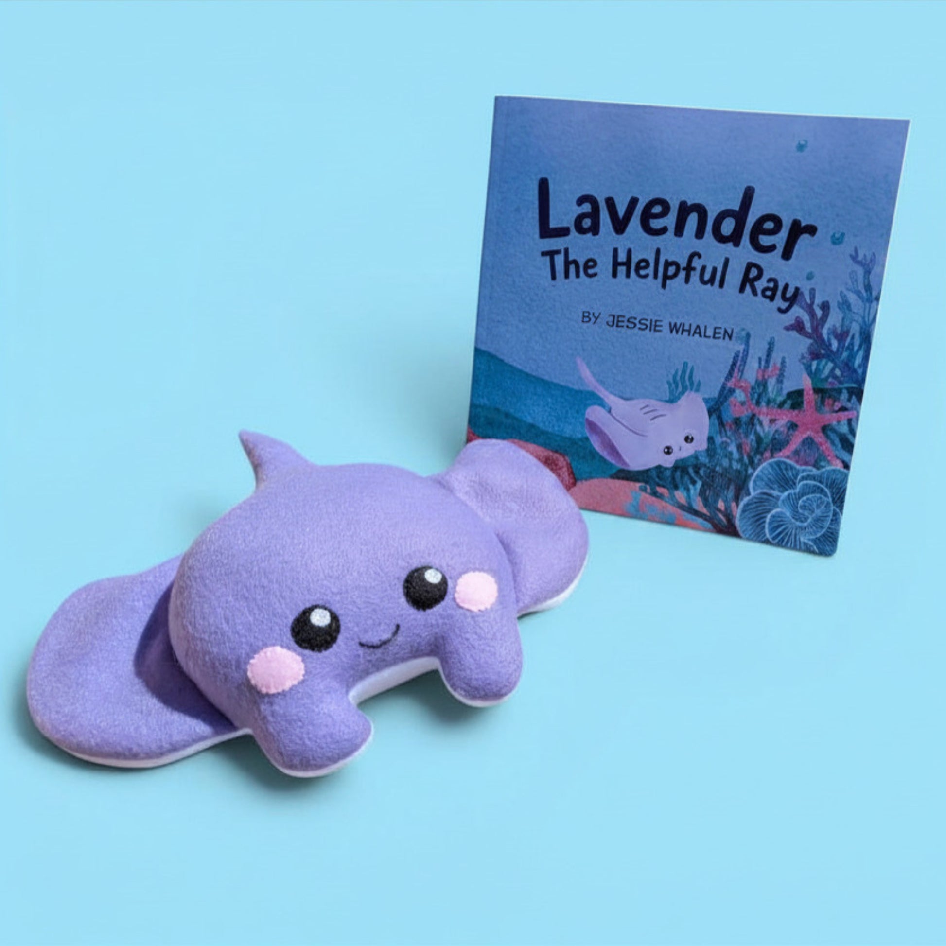 A lavender-purple plush sea creature toy with a smiling face and pink cheeks, placed in front of a children's book titled 'Lavender The Helpful Ray' by Jessie Whalen on a light blue background.