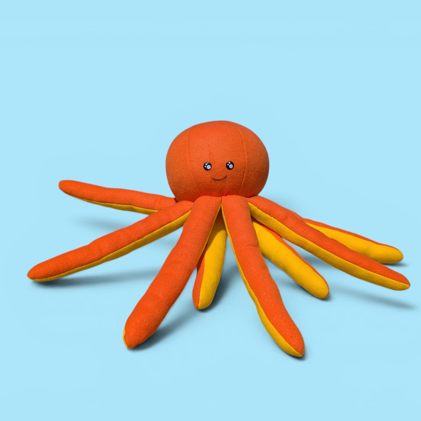 A plush octopus toy with a round orange head, black safety eyes, and a smiling mouth, with long orange and yellow tentacles, shown on a light blue background.