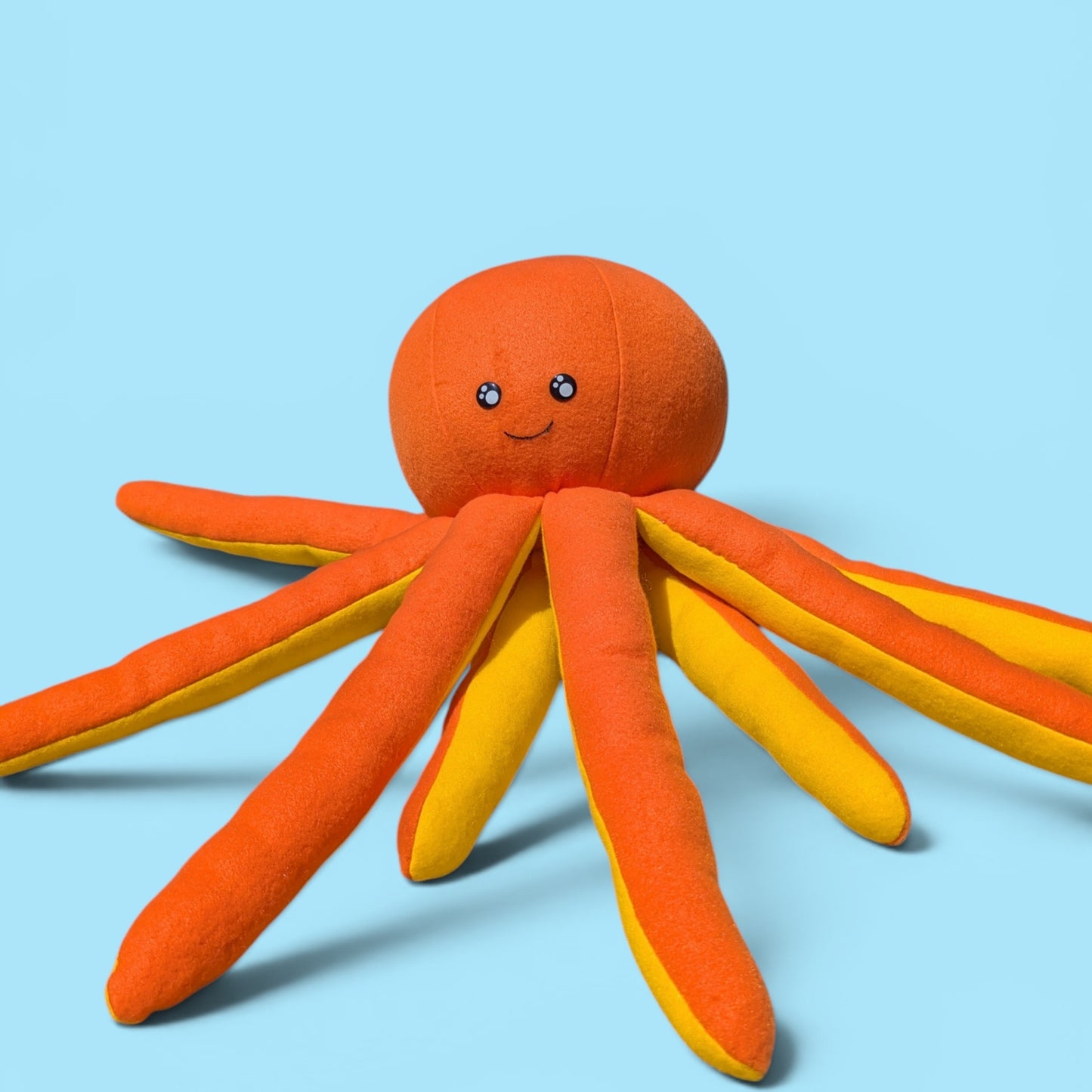 A plush octopus toy with a round orange head, black safety eyes on a smiling face, and eight tentacles that are orange with yellow inner edges, set against a solid light blue background.