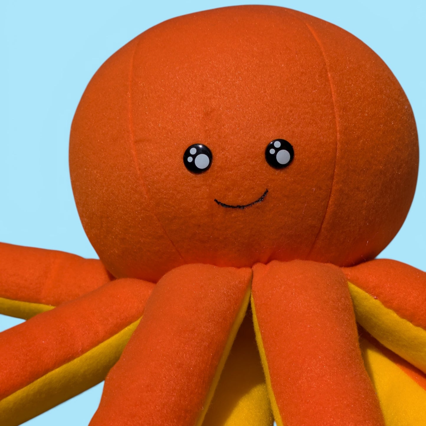 A close-up of a plush octopus toy with a round orange body, eight orange tentacles with yellow inner edges, and a simple face on a light blue background.