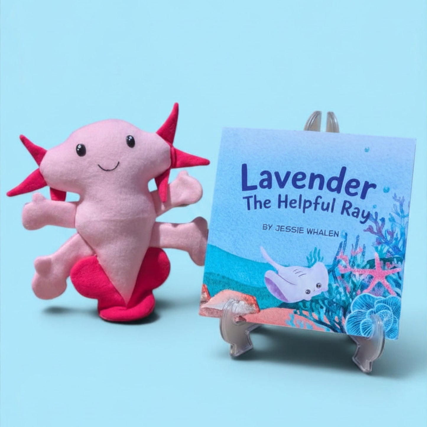 A pink axolotl plush toy sits beside a children's book titled "Lavender The Helpful Ray" by Jessie Whalen, displayed on a small clear stand against a light blue background.