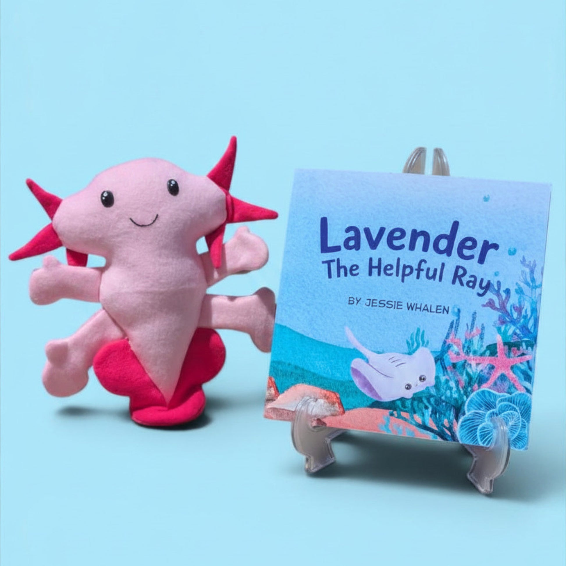 A pink axolotl plush toy sits beside a children's book titled "Lavender The Helpful Ray" by Jessie Whalen, displayed on a small clear stand against a light blue background.