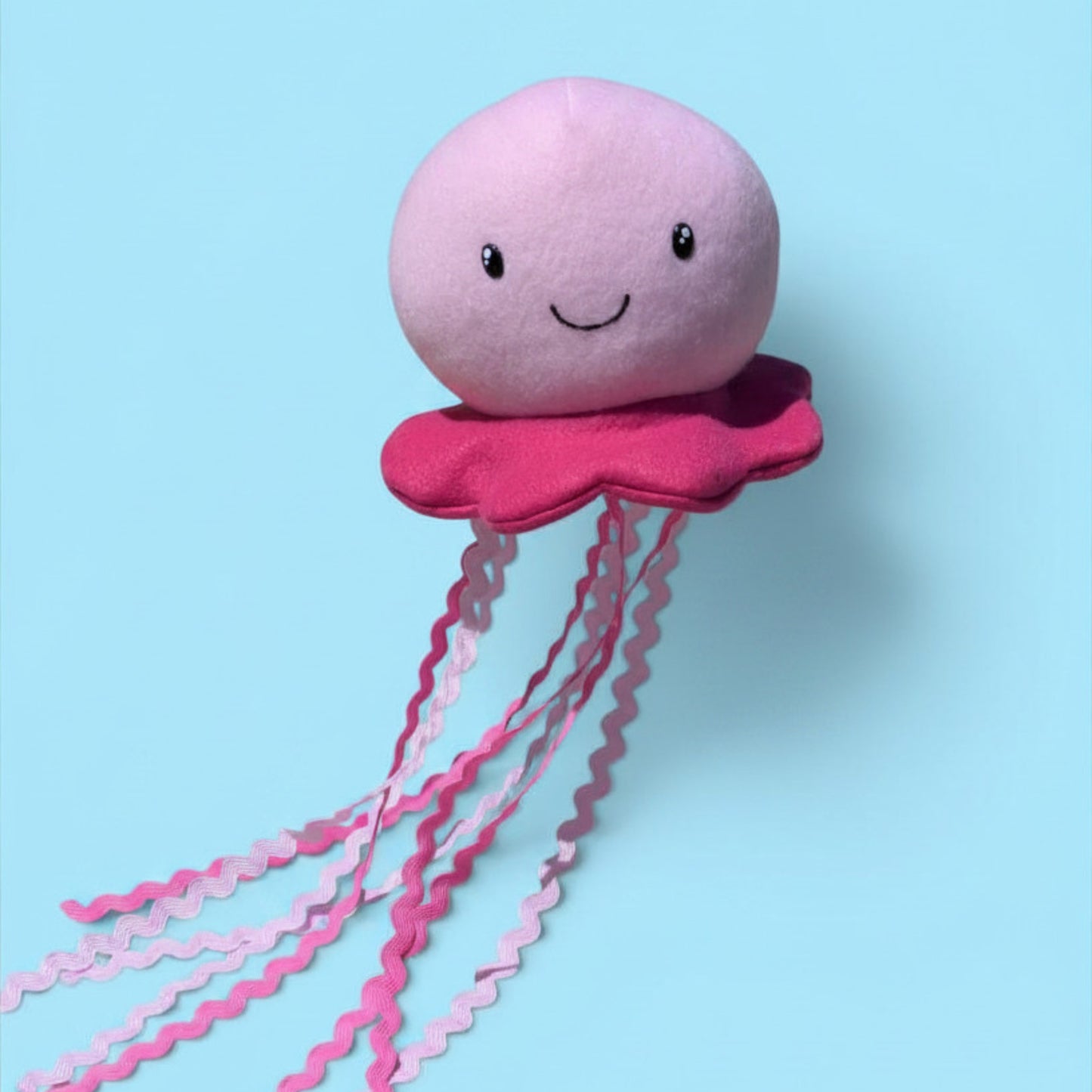 A pink plush jellyfish toy with a smiling face, a darker pink flared bottom, and long wavy pink tentacles, shown against a light blue background.